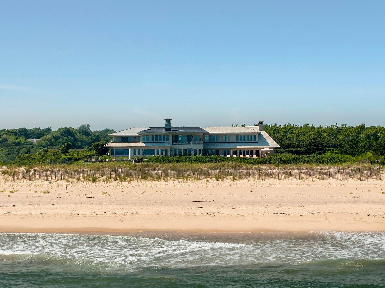 This expansive, multi-level coastal residence is captured from the perspective of the ocean, showcasing its prominent position behind a sandy dune and beach grass. The home features a classic shingle-style aesthetic with a sprawling layout, numerous windows, and a prominent chimney stack. The setting is serene and private, emphasizing the property's direct access to the shoreline and its integration into the natural landscape.
