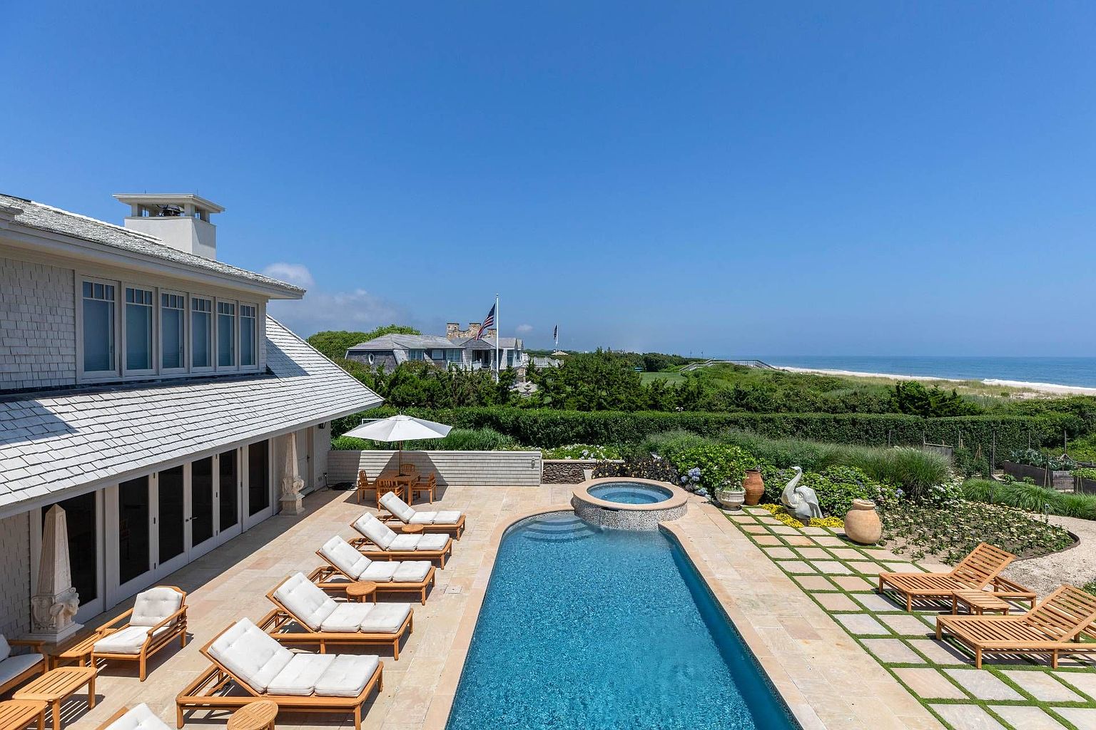 This elevated perspective captures a luxurious coastal backyard featuring a rectangular swimming pool with an integrated circular spa. A stone-paved patio is lined with multiple lounge chairs, while a grid-patterned walkway leads toward a lush garden area overlooking the ocean. The scene is framed by the side of a shingled house with large windows and a clear blue sky, creating a serene and high-end resort-like atmosphere.