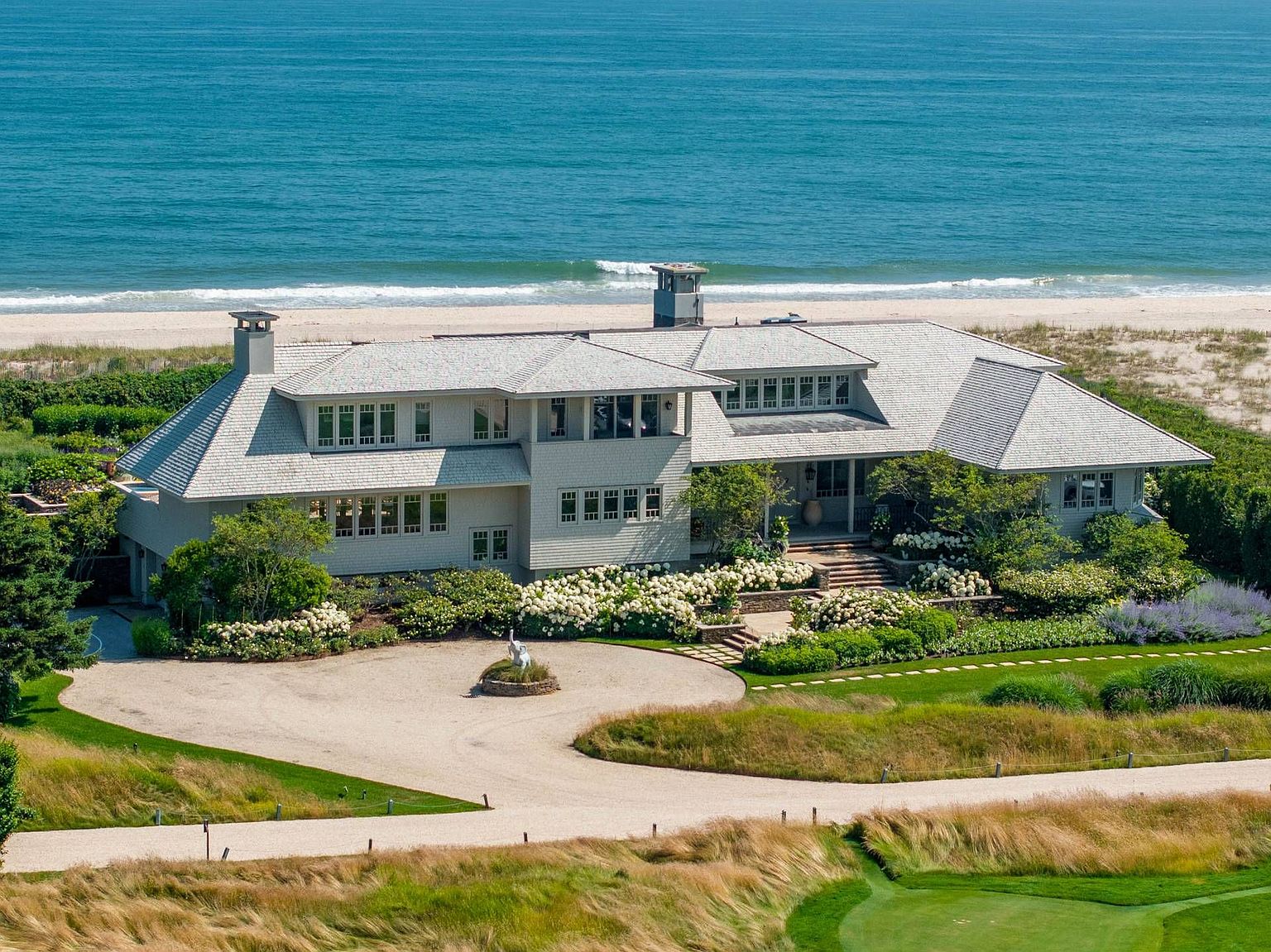 This high-angle aerial view captures a sprawling, shingle-style coastal estate nestled directly against a sandy beach and the ocean. The home features a complex roofline, light-colored siding, and a circular gravel driveway centered around a decorative sculpture, all surrounded by lush, manicured landscaping with abundant white hydrangea bushes. The perspective emphasizes the property's prime waterfront location and its seamless integration into the natural dune landscape.