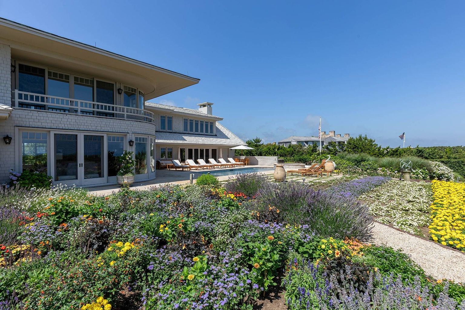 This expansive rear view of a coastal-style residence showcases a multi-level home with light-colored shingle siding and a curved architectural design. The property features a spacious stone patio surrounding a swimming pool, complemented by rows of lounge chairs and a lush, vibrant garden filled with lavender and yellow flowers in the foreground. The scene is captured under a clear blue sky, emphasizing the serene and luxurious outdoor living space.