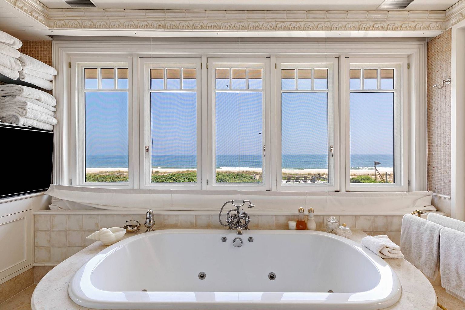 This primary bathroom features a large, oval jetted soaking tub centered beneath a expansive bank of windows that frame a direct, serene view of the ocean and beach. The space is finished with neutral-toned stone surfaces, elegant crown molding, and a stack of plush white towels to the left. The perspective is a straight-on, symmetrical shot that emphasizes the tranquil, spa-like atmosphere and the premium coastal location.