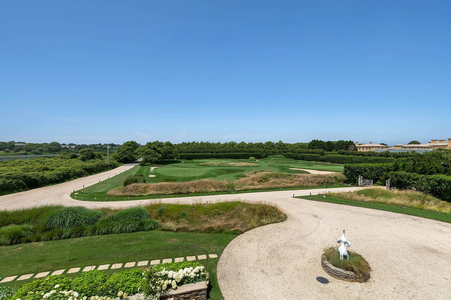 This elevated view captures a sprawling, meticulously landscaped estate grounds featuring a private putting green nestled among rolling dunes and manicured lawns. A winding gravel driveway curves through the property, leading past a decorative sculpture set within a small stone-bordered garden bed. The scene is framed by lush, mature hedges and trees, creating a serene and expansive outdoor retreat under a clear blue sky.