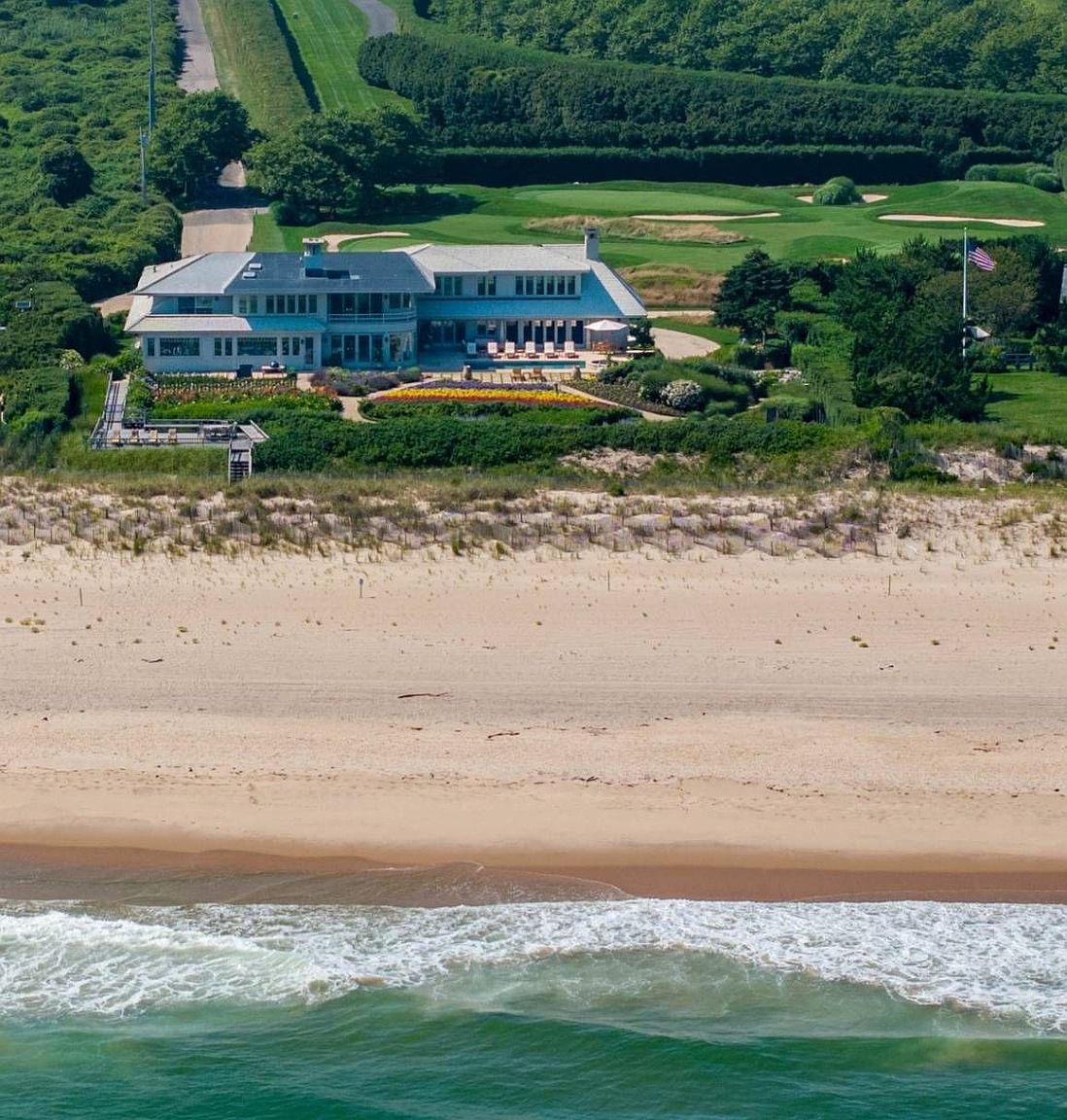 This high-angle aerial shot captures a sprawling coastal estate situated directly on the dunes overlooking the ocean. The property features a large, multi-level residence with a swimming pool, vibrant flower gardens, and a private walkway leading toward the beach. Beyond the house, a manicured golf course stretches across the landscape, creating a seamless blend of luxury living and natural coastal beauty.