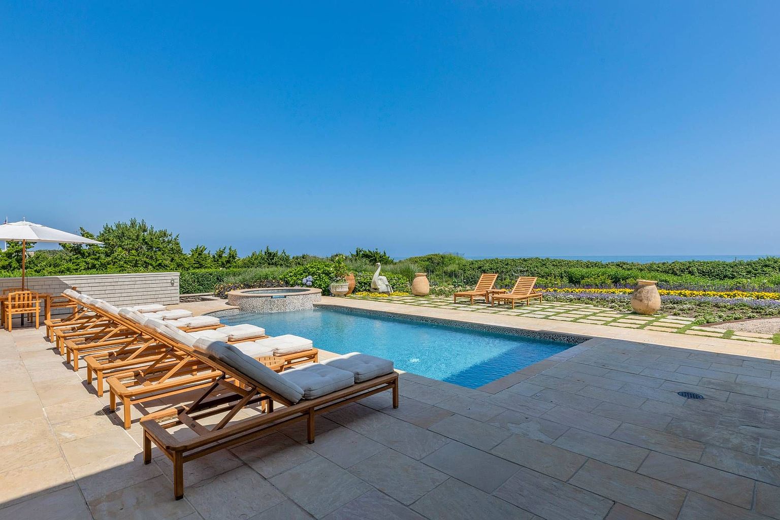 This expansive outdoor living space features a rectangular swimming pool and an integrated circular spa, set against a backdrop of lush greenery and a clear blue ocean horizon. A stone-paved patio surrounds the pool, lined with multiple wooden lounge chairs and a white umbrella, creating a luxurious resort-like atmosphere. The scene is captured from a ground-level perspective, emphasizing the seamless transition between the manicured pool deck and the natural coastal landscape.