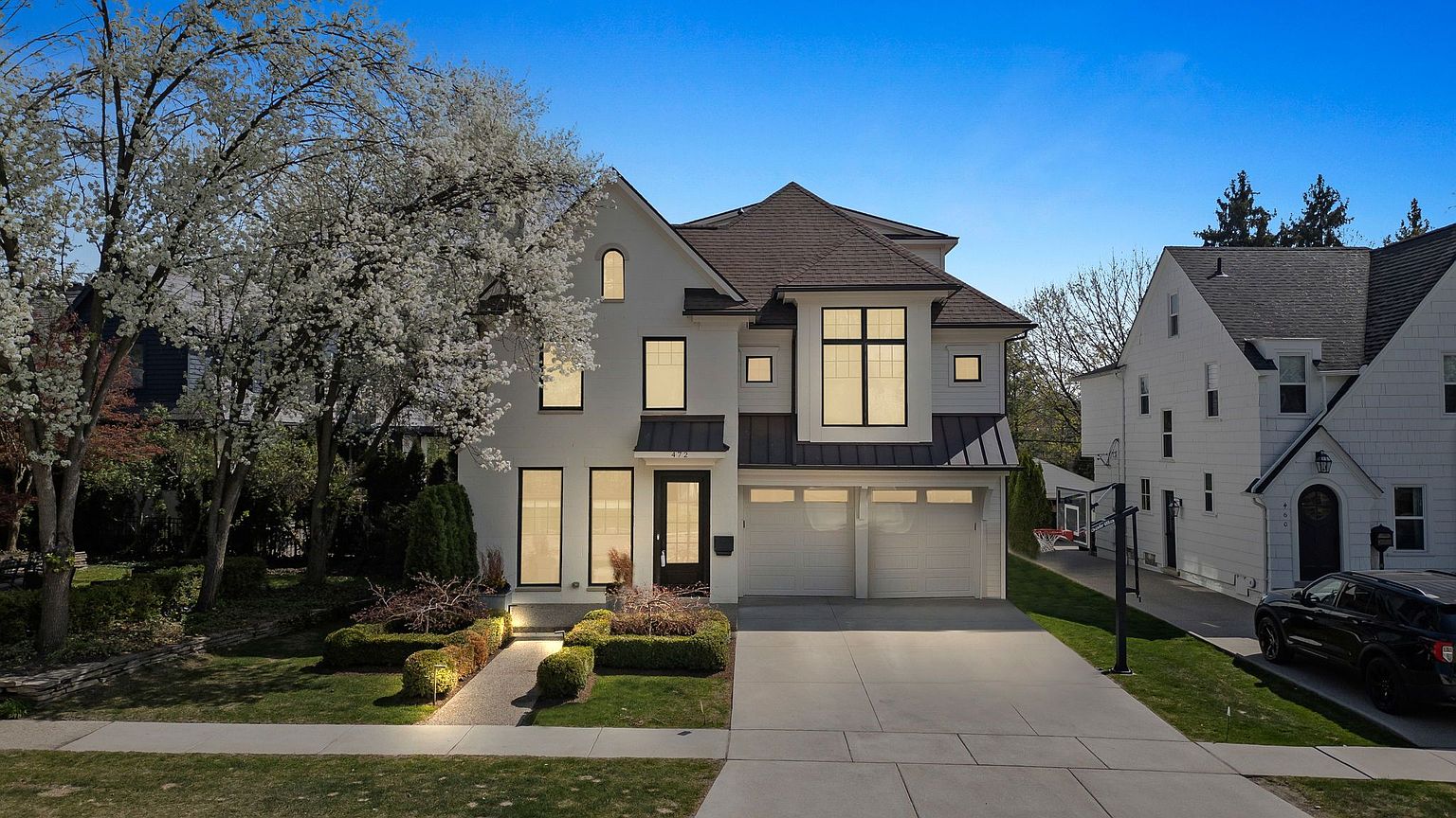 This is a front exterior view of a two-story modern home with a white facade, black-framed windows, and a dark roof. The property features a well-manicured lawn, trimmed hedges, and a paved driveway leading to a two-car garage. A mature tree with white blossoms adds to the curb appeal, creating an inviting and sophisticated impression.