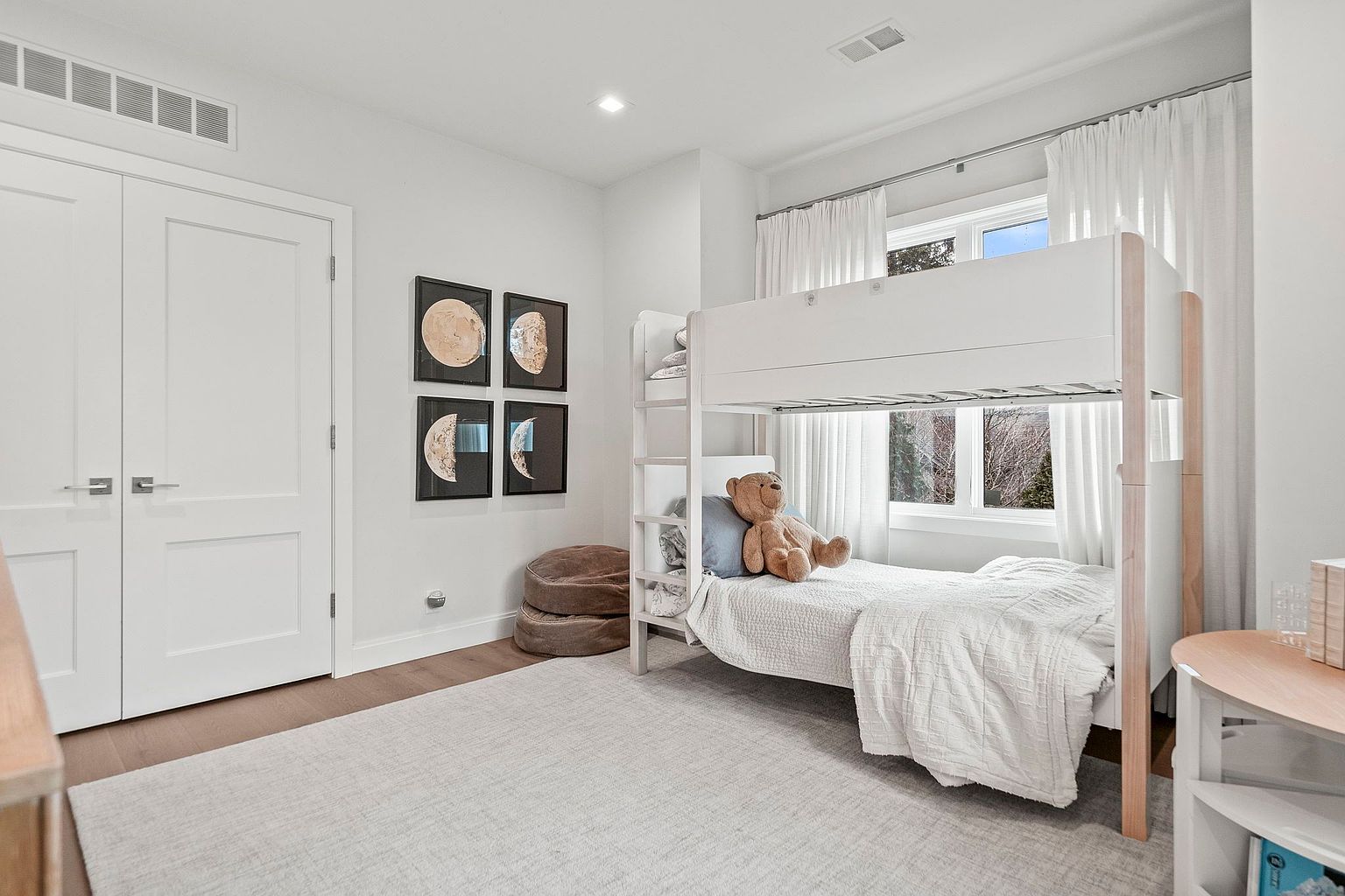 This is a bright and airy bedroom featuring a white bunk bed with a teddy bear on the lower bunk. The room is decorated with moon phase artwork and neutral tones, creating a calming atmosphere. Natural light streams in through the window, complemented by sheer white curtains.
