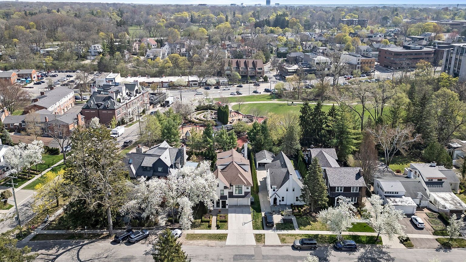 This aerial view showcases a charming residential neighborhood with well-maintained homes, mature trees, and a community park. The houses feature a mix of architectural styles, with manicured lawns and blooming trees adding to the curb appeal. The overall impression is one of a peaceful and desirable place to live.