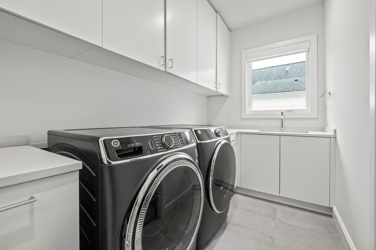 This is a modern laundry room featuring sleek, dark-colored front-loading washer and dryer units. White cabinetry above and to the side provides ample storage space, complemented by a bright window over a sink area. The room has a clean and minimalist aesthetic, perfect for a contemporary home.