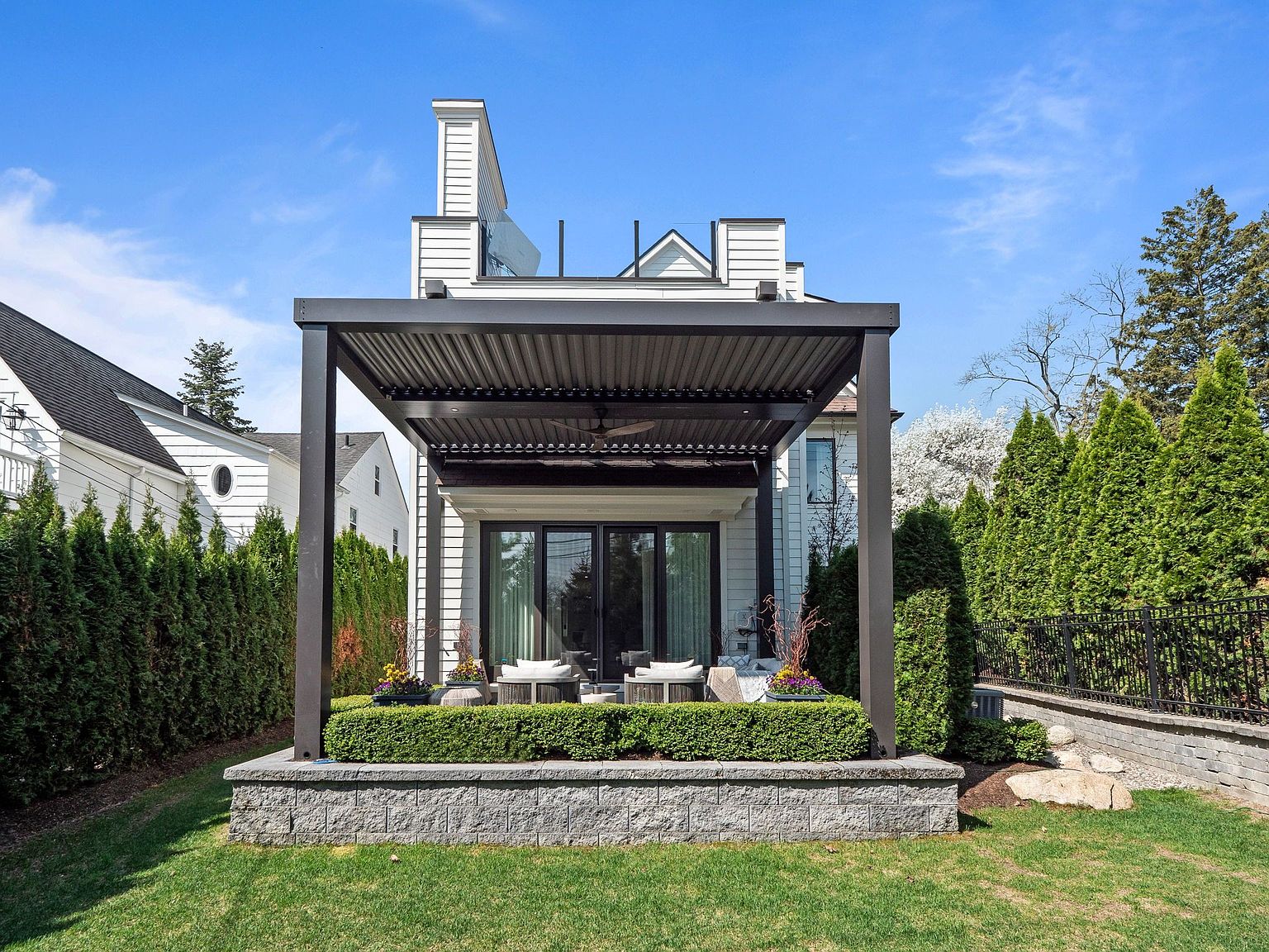 This image showcases a beautifully designed outdoor patio area. The patio features a modern pergola with a dark frame, providing shade and a stylish focal point. The space is elevated on a stone platform, surrounded by manicured greenery, creating a private and inviting outdoor living area.