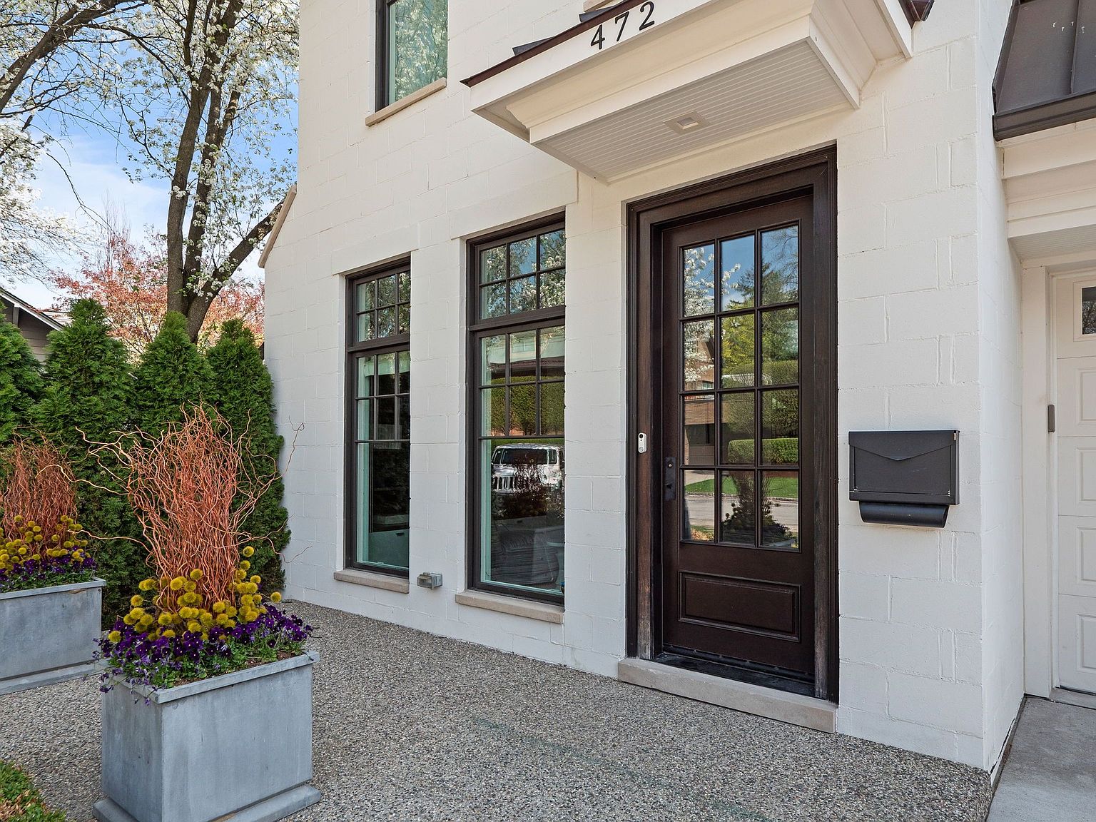 This is a front exterior shot of a home's entryway, showcasing a dark wood front door with multi-pane glass, flanked by two windows with dark frames. The house is painted white brick, and the address '472' is visible above the door. Planters with colorful flowers and greenery add a touch of nature to the entrance, creating a welcoming and stylish curb appeal.