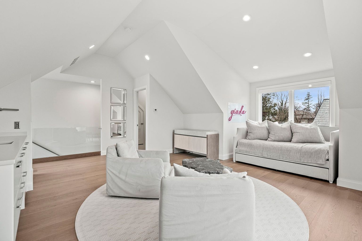 This is a bright and airy living room featuring a neutral color palette with white walls and light wood floors. The room is furnished with comfortable seating, including two armchairs and a daybed, arranged around a round rug and a unique coffee table. Natural light floods the space through a large window, creating a welcoming and serene atmosphere.