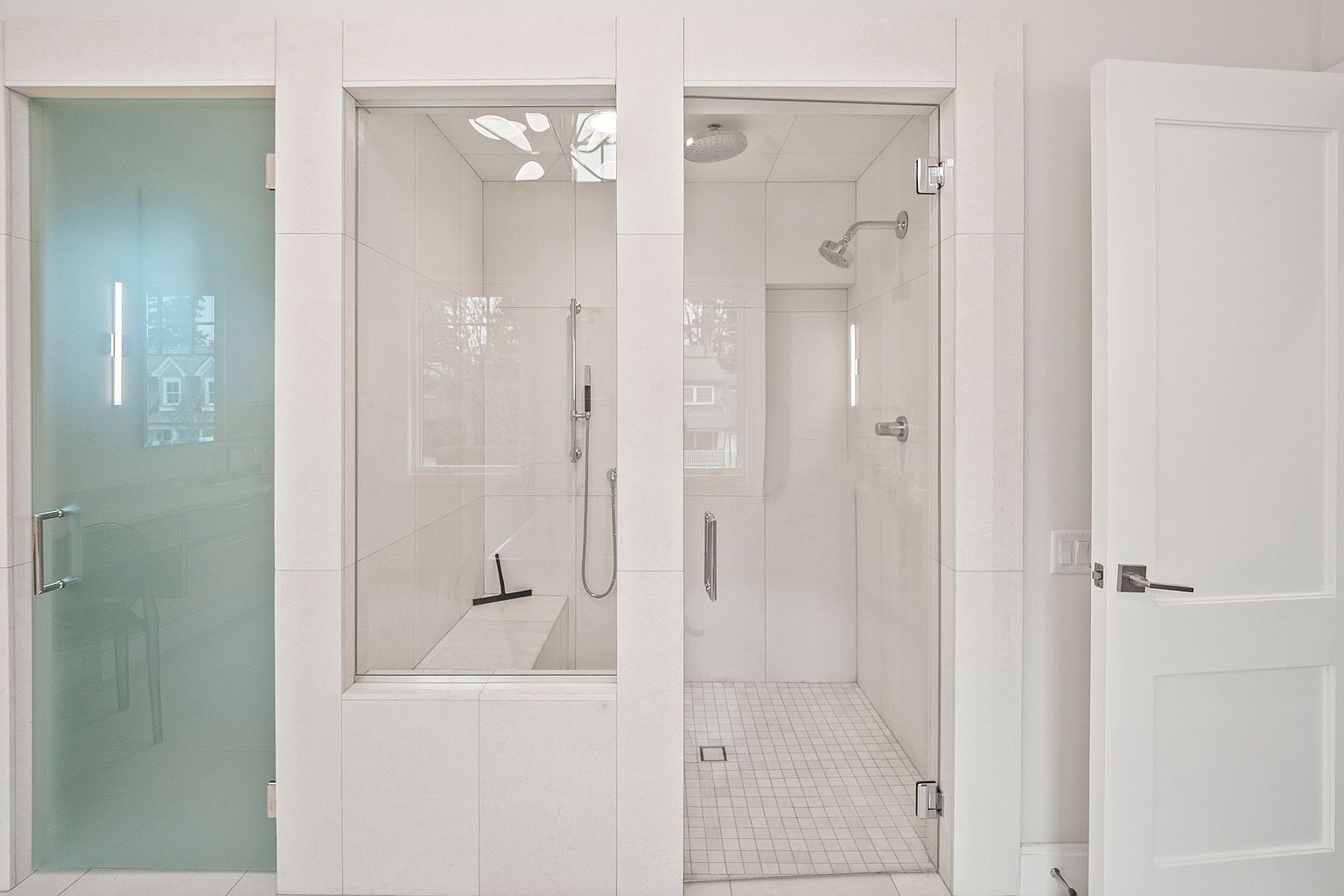This is a bright and modern primary bathroom featuring two glass-enclosed showers with white tile and chrome fixtures. The left shower has a frosted glass door, while the right shower has a clear glass door. A white door is visible on the right side of the image, and the overall impression is clean and luxurious.