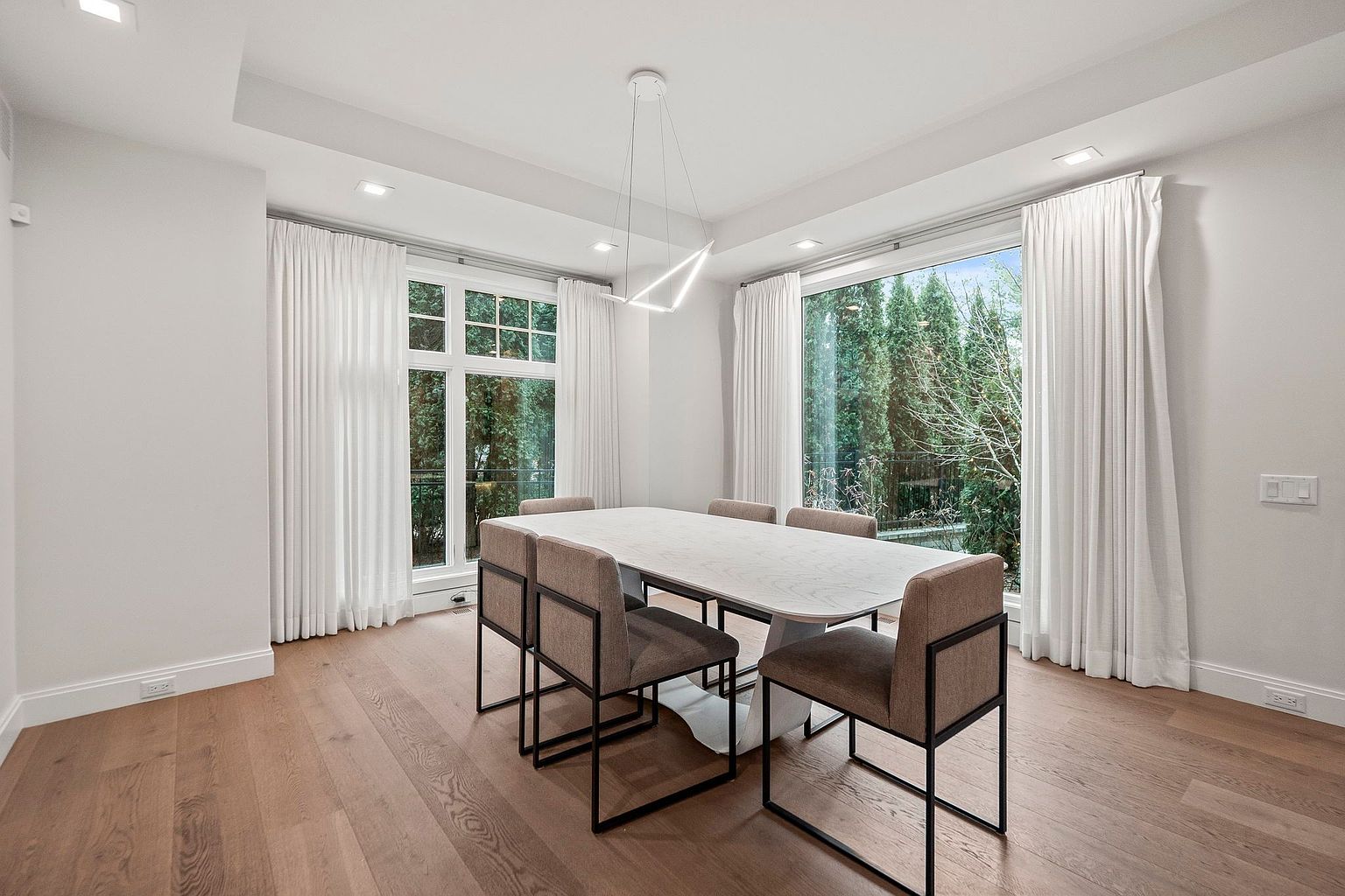 This is a bright and airy dining room featuring a modern aesthetic. A rectangular dining table with a light-colored top is surrounded by six chairs with dark metal frames and neutral-toned upholstery. Large windows with white curtains provide ample natural light and views of the greenery outside, complemented by a contemporary geometric chandelier.