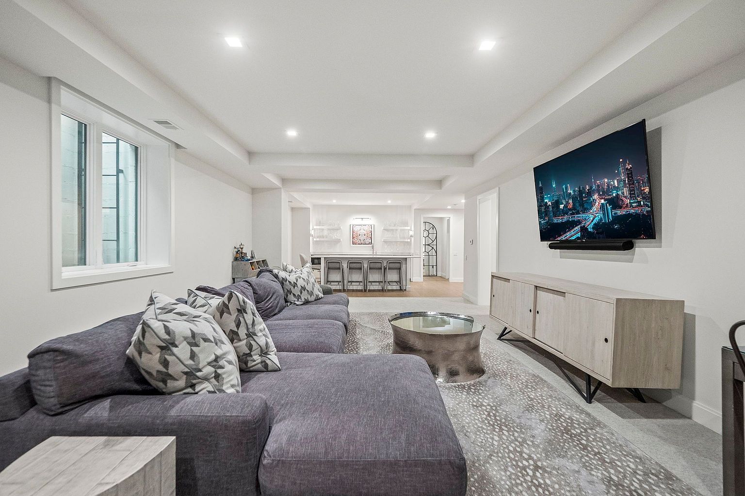 This is a well-lit, modern living room featuring a large, comfortable sectional sofa in a neutral tone, complemented by decorative pillows. A sleek, low-profile media console sits beneath a large flat-screen TV displaying a cityscape at night. The room also includes a unique coffee table and a stylish rug, creating a cozy and inviting atmosphere.