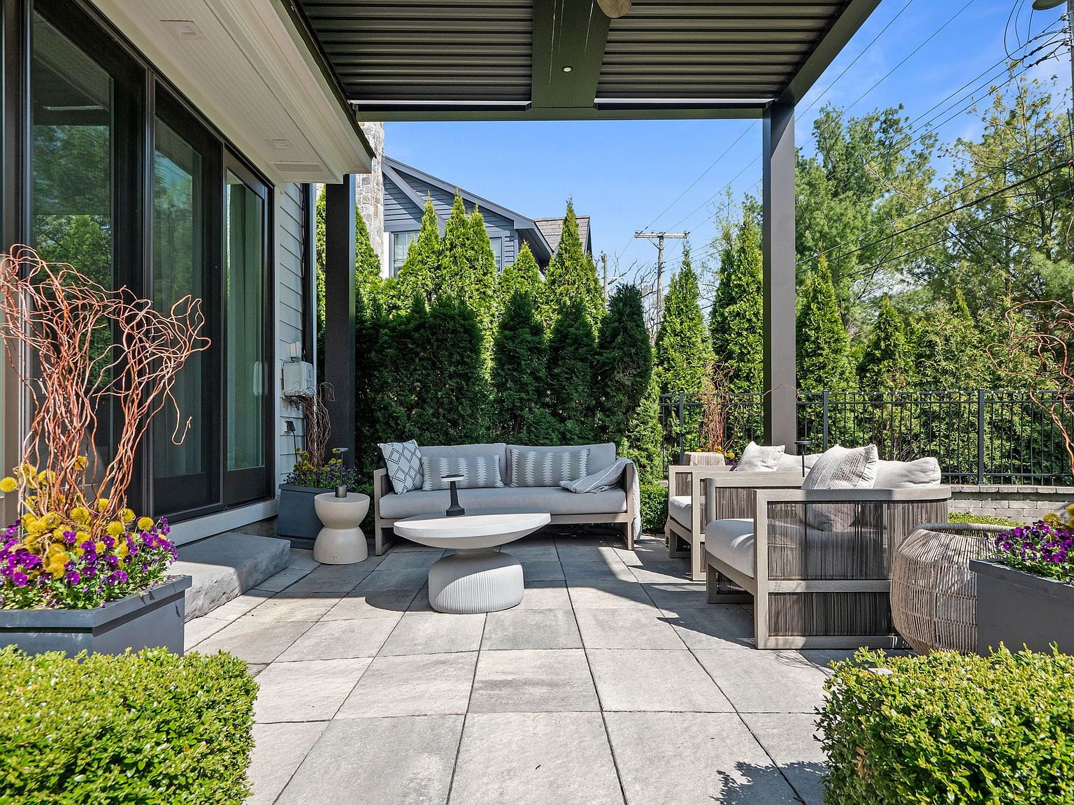 This image showcases a stylish outdoor patio area, featuring modern furniture including a sofa, chairs, and a unique round coffee table. The patio is paved with large square tiles and is partially covered by a dark-colored pergola, providing shade. Lush greenery and potted plants add a touch of nature and privacy to the space, creating an inviting and relaxing atmosphere.