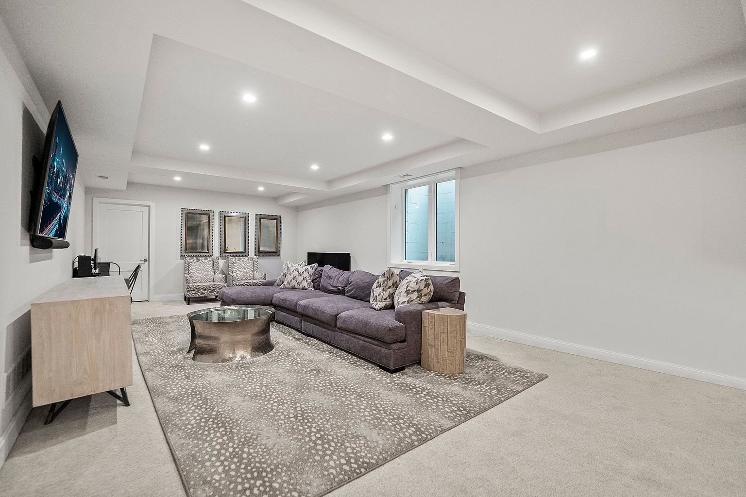 This is an interior shot of a modern living room. The room features a large gray sectional sofa, a unique metal coffee table, and a patterned area rug. A flat-screen TV is mounted on the wall, and natural light enters through a window, creating a bright and inviting space.