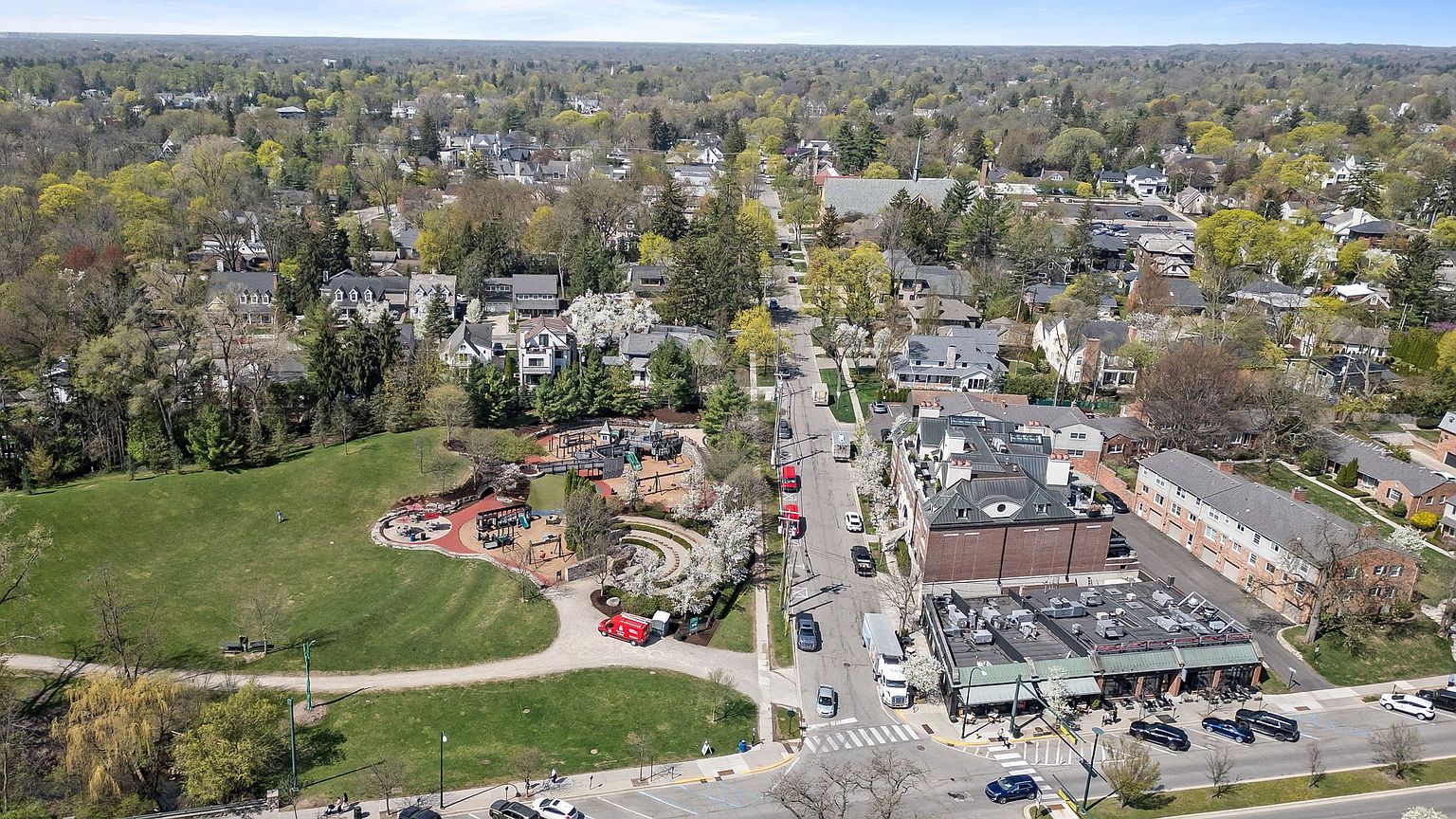This aerial view showcases a charming neighborhood with a mix of residential homes, mature trees, and a well-maintained park. The park features a playground and walking paths, adding to the community's appeal. A commercial area with a brick building and outdoor seating is also visible, suggesting convenient access to local amenities.
