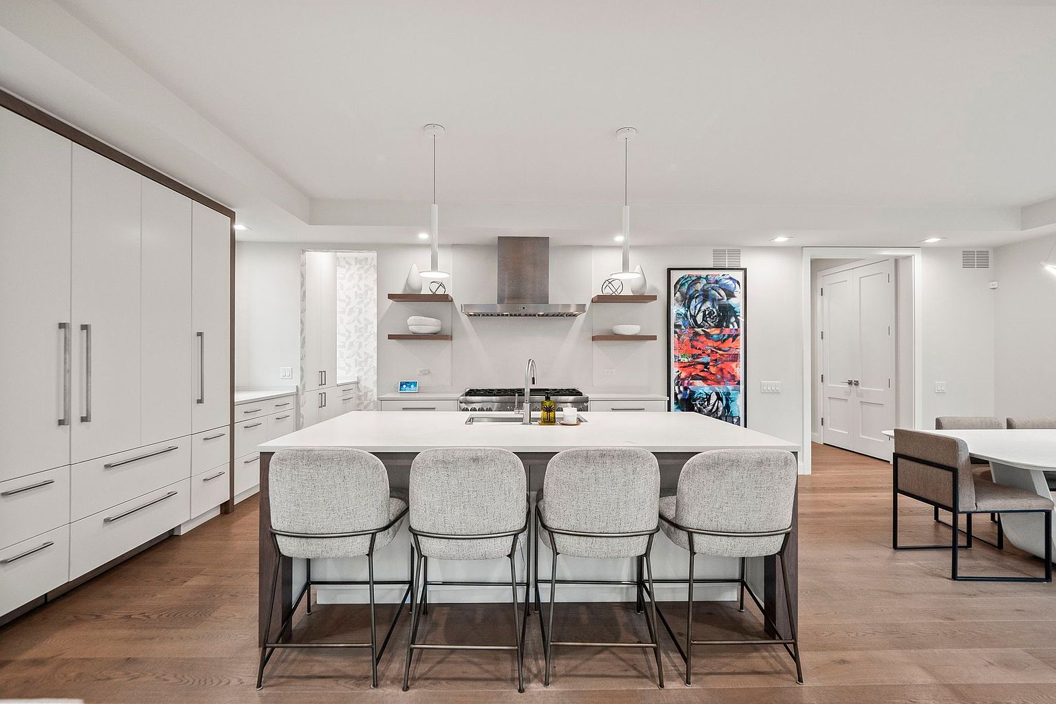 This is a bright and modern kitchen featuring a large central island with seating, white cabinetry with sleek hardware, and stainless steel appliances. The open layout connects to a dining area, and the hardwood flooring adds warmth to the space. The kitchen is well-lit with pendant lights and recessed lighting, creating an inviting atmosphere.