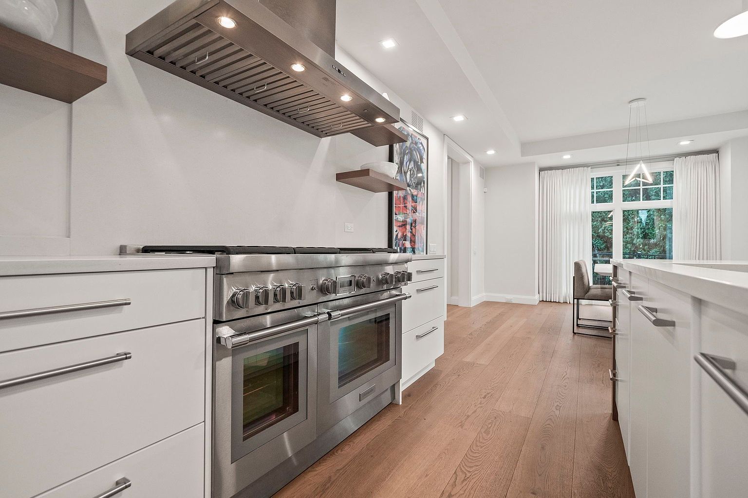 This is a bright and modern kitchen featuring white cabinetry, stainless steel appliances, and hardwood flooring. A large stainless steel range with a matching vent hood is the focal point, complemented by floating shelves and a piece of artwork. The perspective is from a low angle, emphasizing the depth and spaciousness of the kitchen.