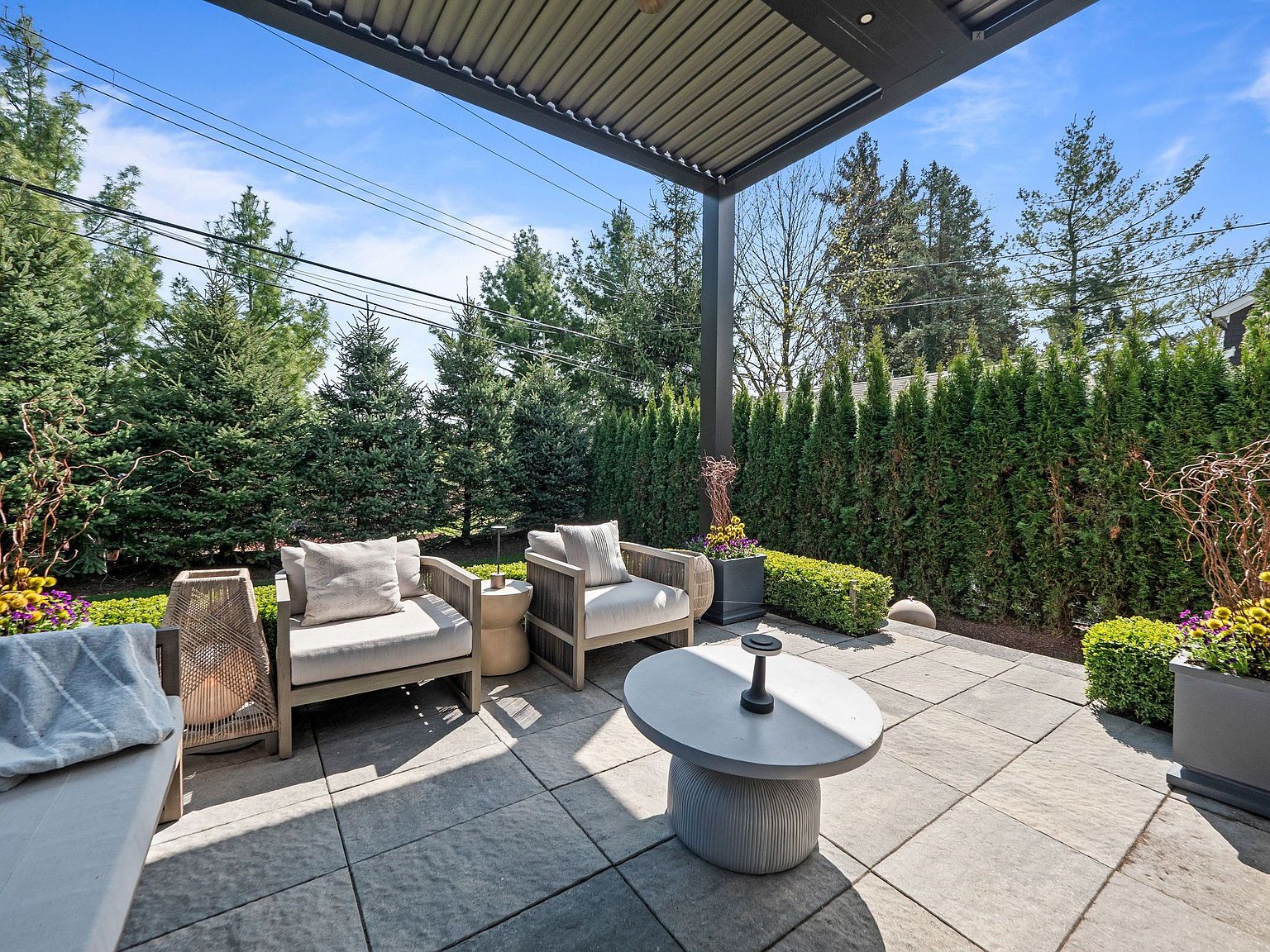 This is a well-appointed outdoor patio area, featuring comfortable seating arrangements with cushioned chairs and a modern round coffee table. The patio is paved with large, square tiles and is partially covered by a pergola, providing shade. Lush greenery, including tall hedges and potted plants, surrounds the space, creating a private and serene atmosphere.