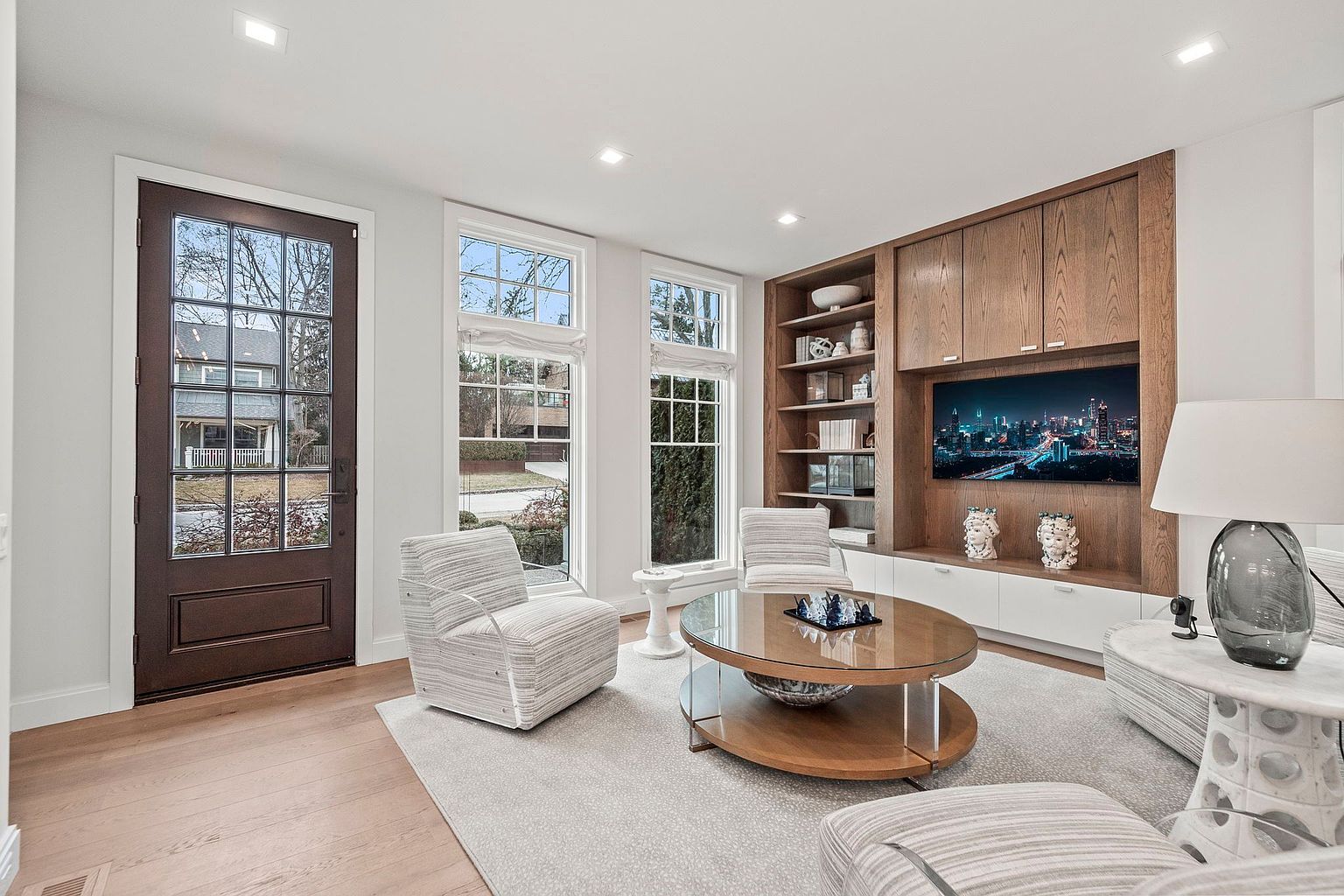This is an interior shot of a living room featuring a modern design with a focus on natural light and neutral tones. The room includes a dark wood front door, several windows, striped armchairs, a round glass-top coffee table, and a built-in entertainment center with a television. The flooring is light hardwood, and a large area rug anchors the seating arrangement, creating a cozy and inviting atmosphere.