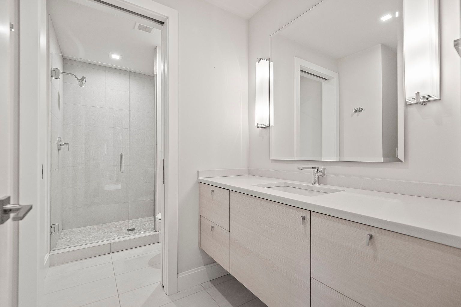 This is a bright and modern primary bathroom featuring a large vanity with light wood-tone cabinets and a white countertop. A frameless mirror reflects the clean lines of the space, complemented by sleek vertical light fixtures. The walk-in shower has glass doors and light gray tile, contributing to the bathroom's minimalist and luxurious feel.