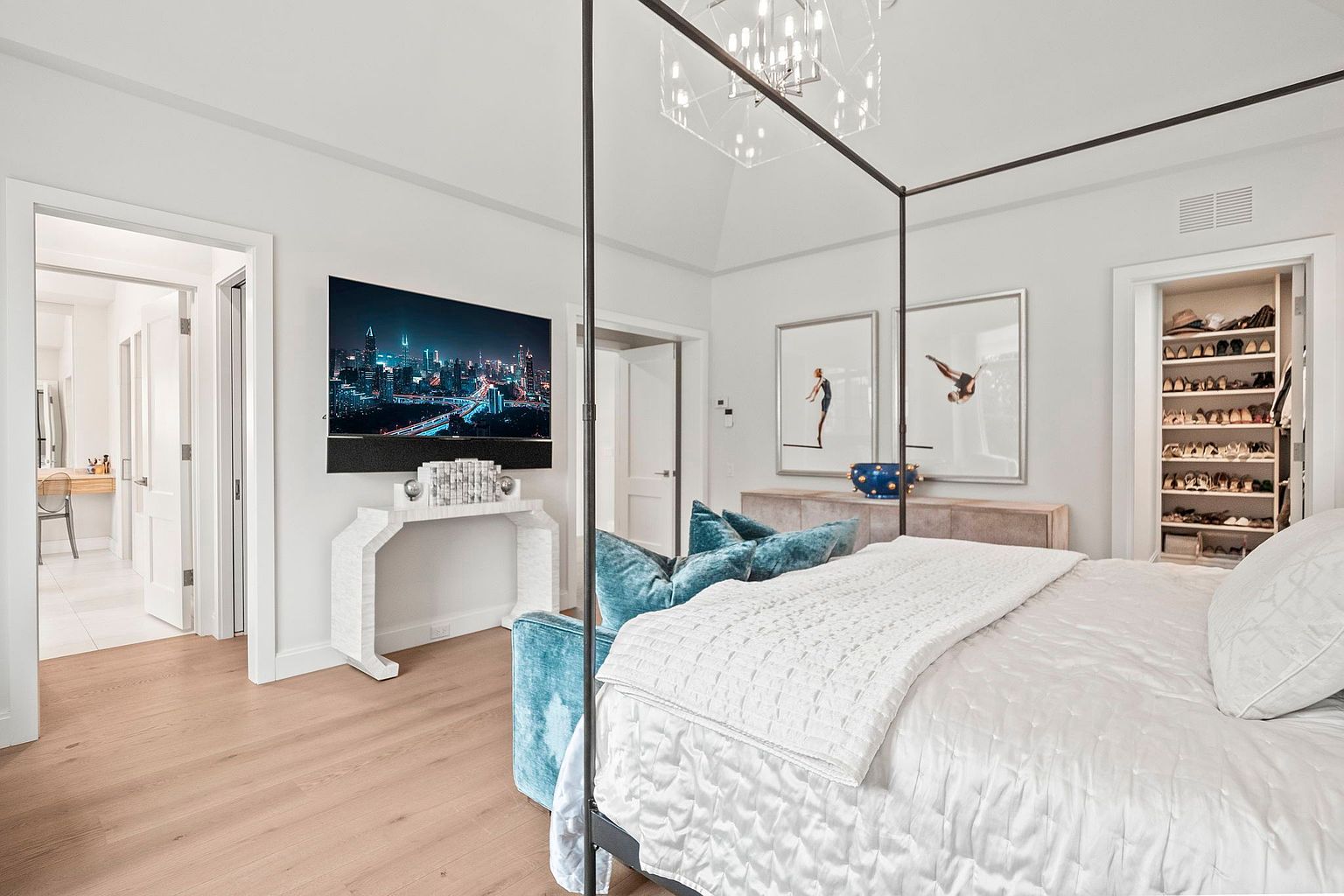 This is a luxurious primary bedroom featuring a four-poster bed with a white quilted comforter and plush blue pillows. A modern TV is mounted above a white console table, and artwork adorns the walls. The room has light hardwood floors and a walk-in closet, creating a serene and upscale atmosphere.