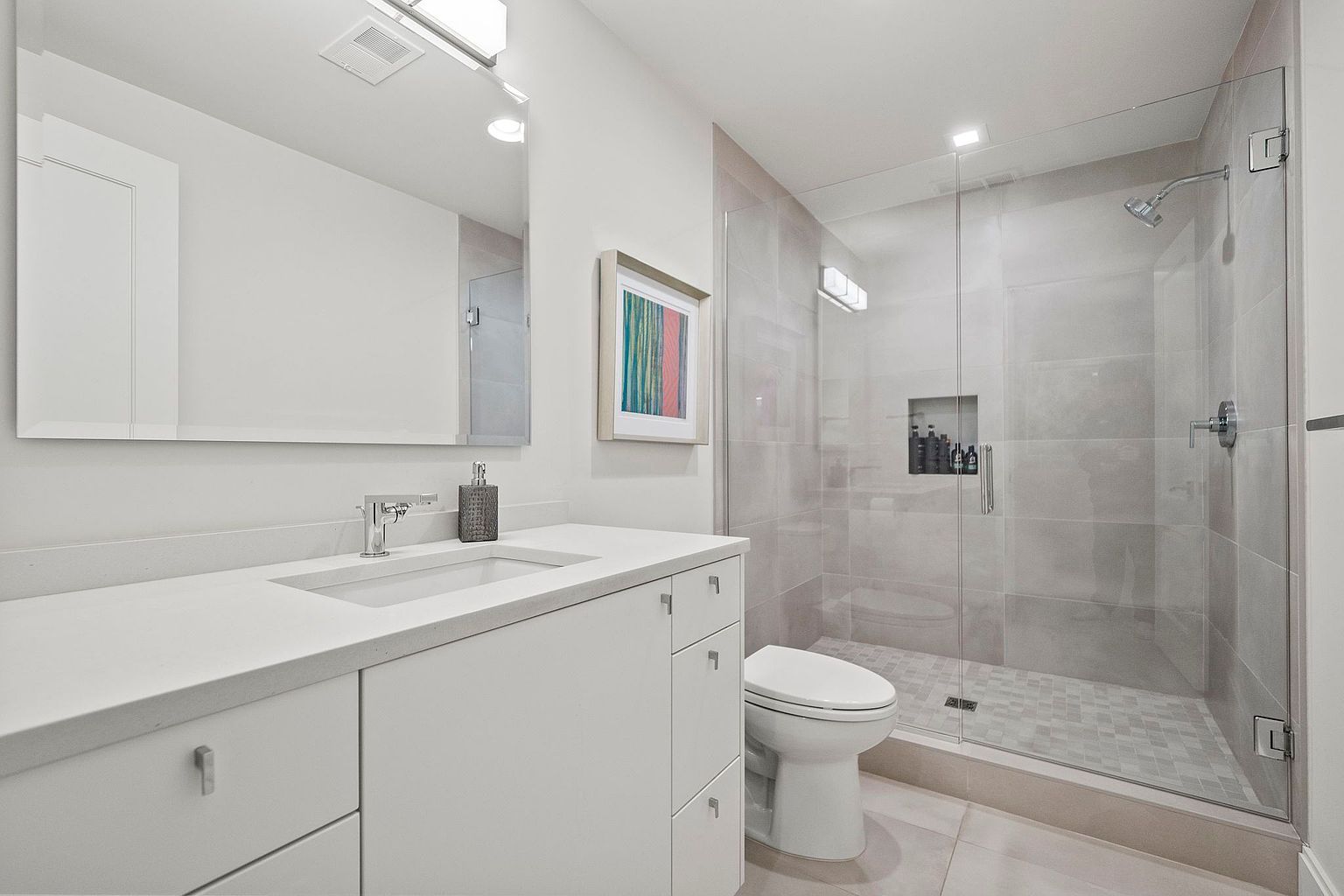 This is a well-lit bathroom featuring a white vanity with a modern sink and faucet, complemented by a large mirror and contemporary lighting fixtures. A glass-enclosed shower with gray tiling and a built-in niche adds a touch of luxury, while a toilet is neatly positioned beside the shower. The overall aesthetic is clean, bright, and minimalist.