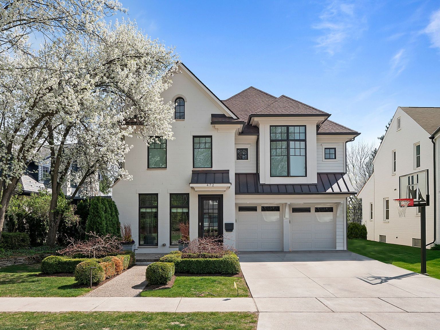 This is a front exterior view of a two-story white brick house with black-framed windows and a brown roof. The house features a two-car garage, manicured landscaping, and a walkway leading to the front door. A large flowering tree adds to the curb appeal, creating an inviting and elegant impression.