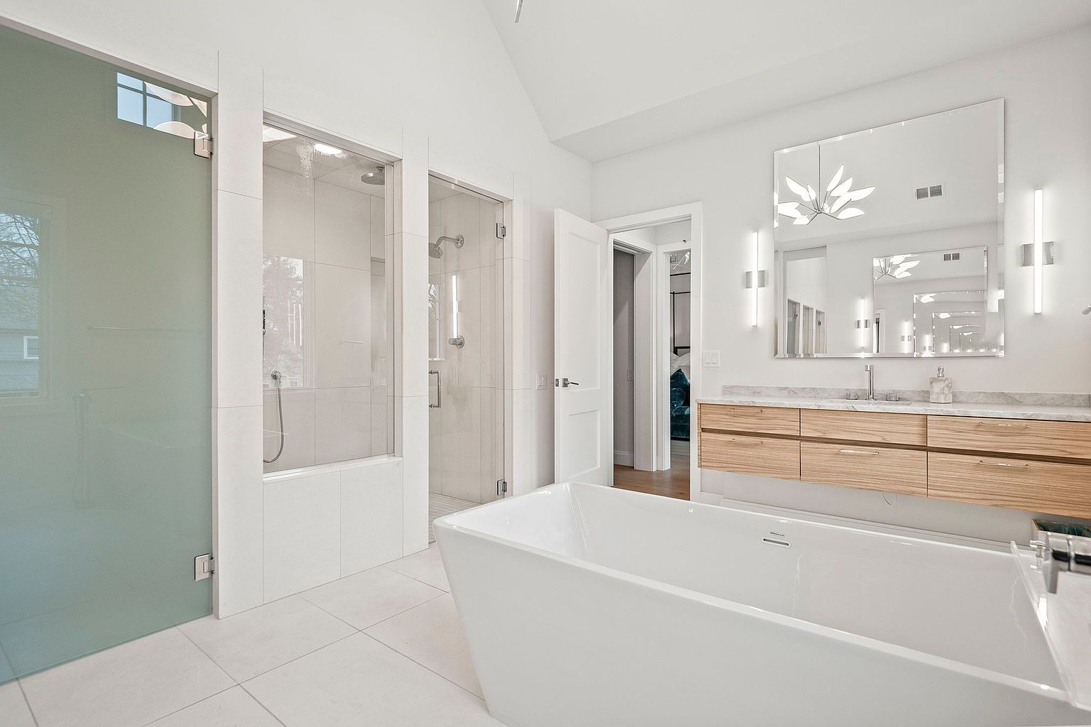 This is a bright and modern primary bathroom featuring a freestanding white bathtub, a double vanity with light wood drawers and a marble countertop, and a large mirror with sconces. The bathroom also includes a glass-enclosed shower and a separate frosted glass shower, all contributing to a clean and luxurious aesthetic. The perspective is from the entrance of the bathroom, showcasing the depth and layout of the space.