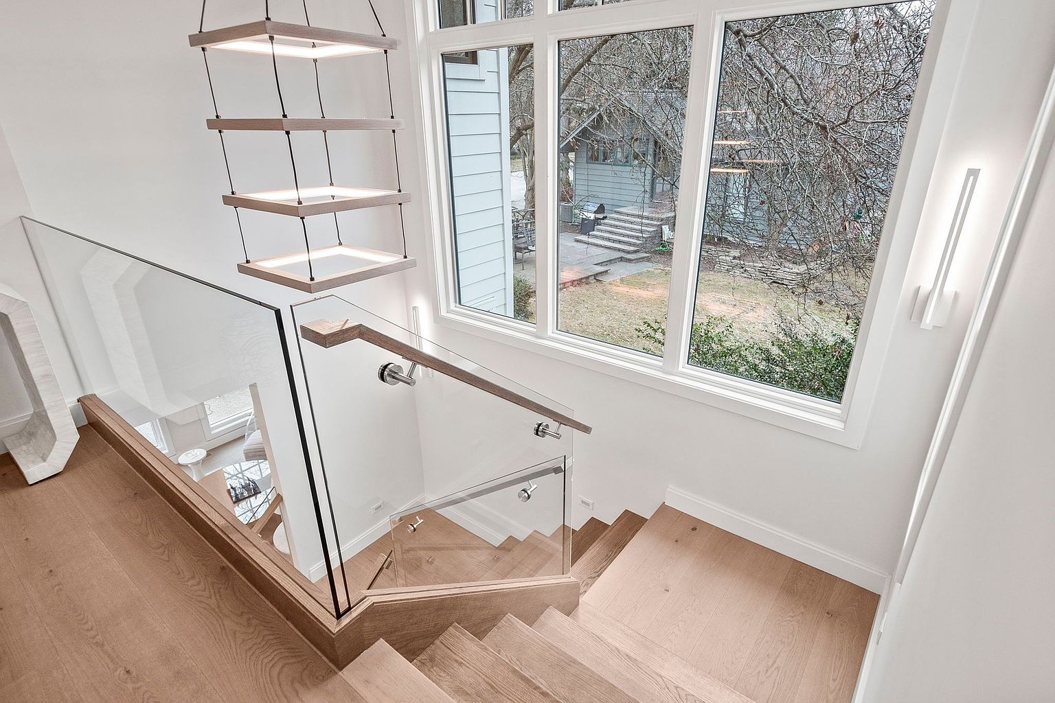 This image showcases a modern staircase with light wood steps and a glass railing, creating an open and airy feel. A unique, multi-tiered pendant light fixture hangs above, complementing the natural light streaming in from a large window overlooking the backyard. The clean lines and minimalist design contribute to a sophisticated and inviting atmosphere.