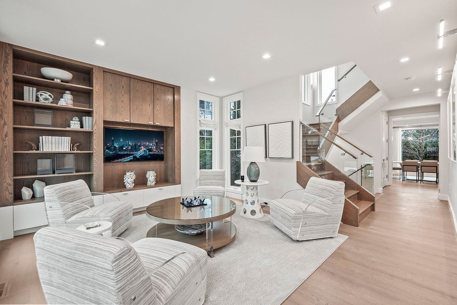 This is a bright and airy living room featuring a built-in entertainment center with wooden shelving and cabinets, a modern TV, and decorative vases. The room is furnished with three striped armchairs arranged around a round glass coffee table on a light-colored rug. A staircase with glass railings is visible in the background, adding to the contemporary feel of the space.