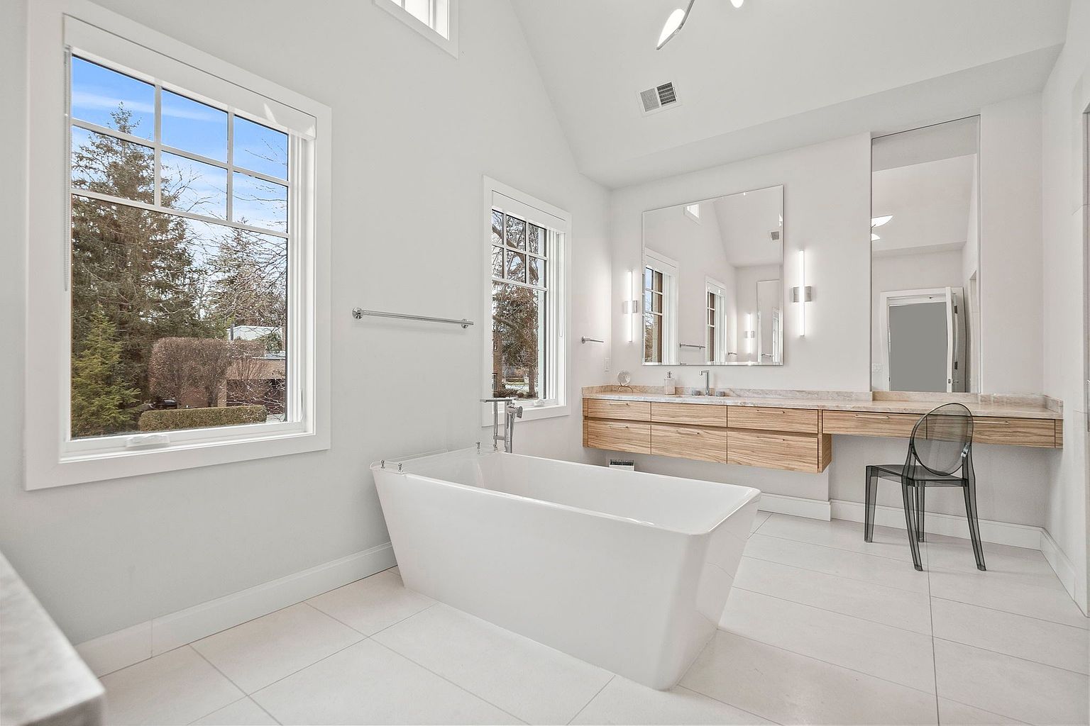 This is a bright and spacious primary bathroom featuring a modern freestanding bathtub positioned near a large window offering natural light and views of the outdoors. The bathroom includes a floating vanity with wood drawers and a light countertop, complemented by a large mirror and a clear acrylic chair. The white tile flooring and walls contribute to the clean and luxurious aesthetic.
