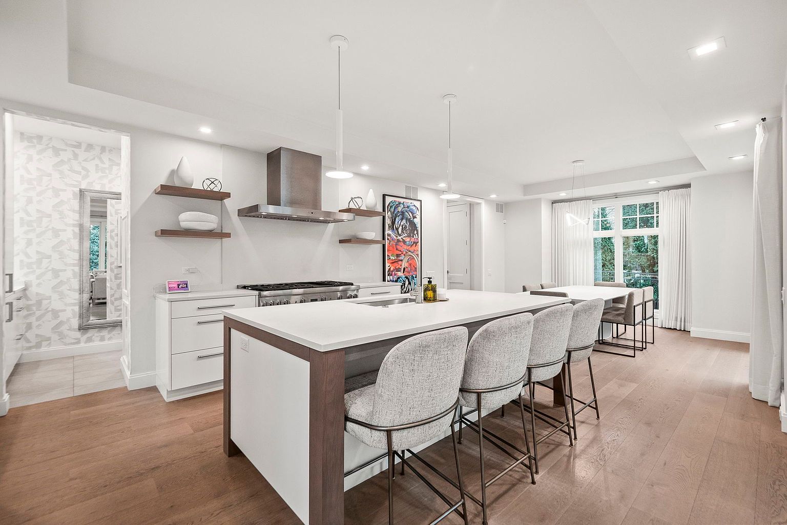 This is a bright and modern kitchen featuring a large island with bar seating, white cabinetry, and stainless steel appliances. The kitchen is well-lit with pendant lighting and natural light from a nearby window. The hardwood floors add warmth to the space, creating an inviting atmosphere.