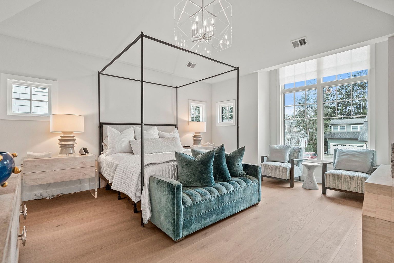 This is a bright and airy primary bedroom featuring a four-poster bed with white linens and a tufted teal velvet bench at the foot. The room is well-lit with natural light from a large window and features neutral tones, creating a serene and luxurious atmosphere. Two armchairs are placed near the window, suggesting a comfortable seating area.