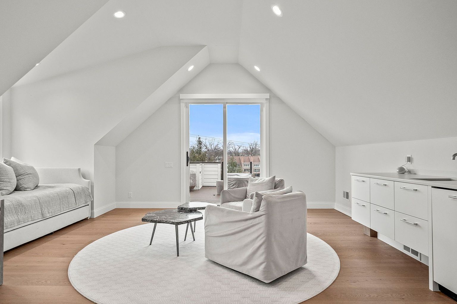 This is an interior shot of a finished attic space, likely intended as a living area. The room features a daybed, two armchairs, a small table, and built-in cabinetry with a sink. A sliding glass door leads to a balcony, and the space is illuminated by recessed lighting, creating a bright and inviting atmosphere.