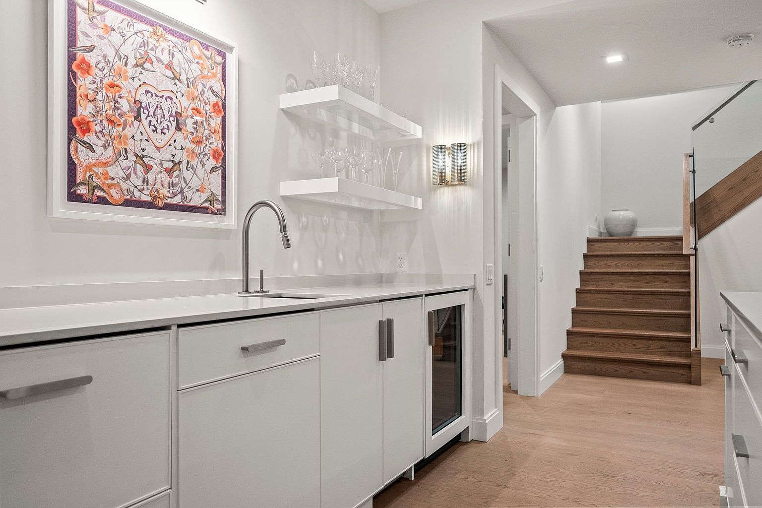 This is a well-lit basement area featuring a stylish wet bar with white cabinetry, a stainless steel sink, and a built-in wine cooler. The bar area is complemented by floating shelves displaying glassware and a decorative wall sconce. Adjacent to the bar, wooden stairs lead to another level, adding architectural interest and suggesting a functional and inviting space.