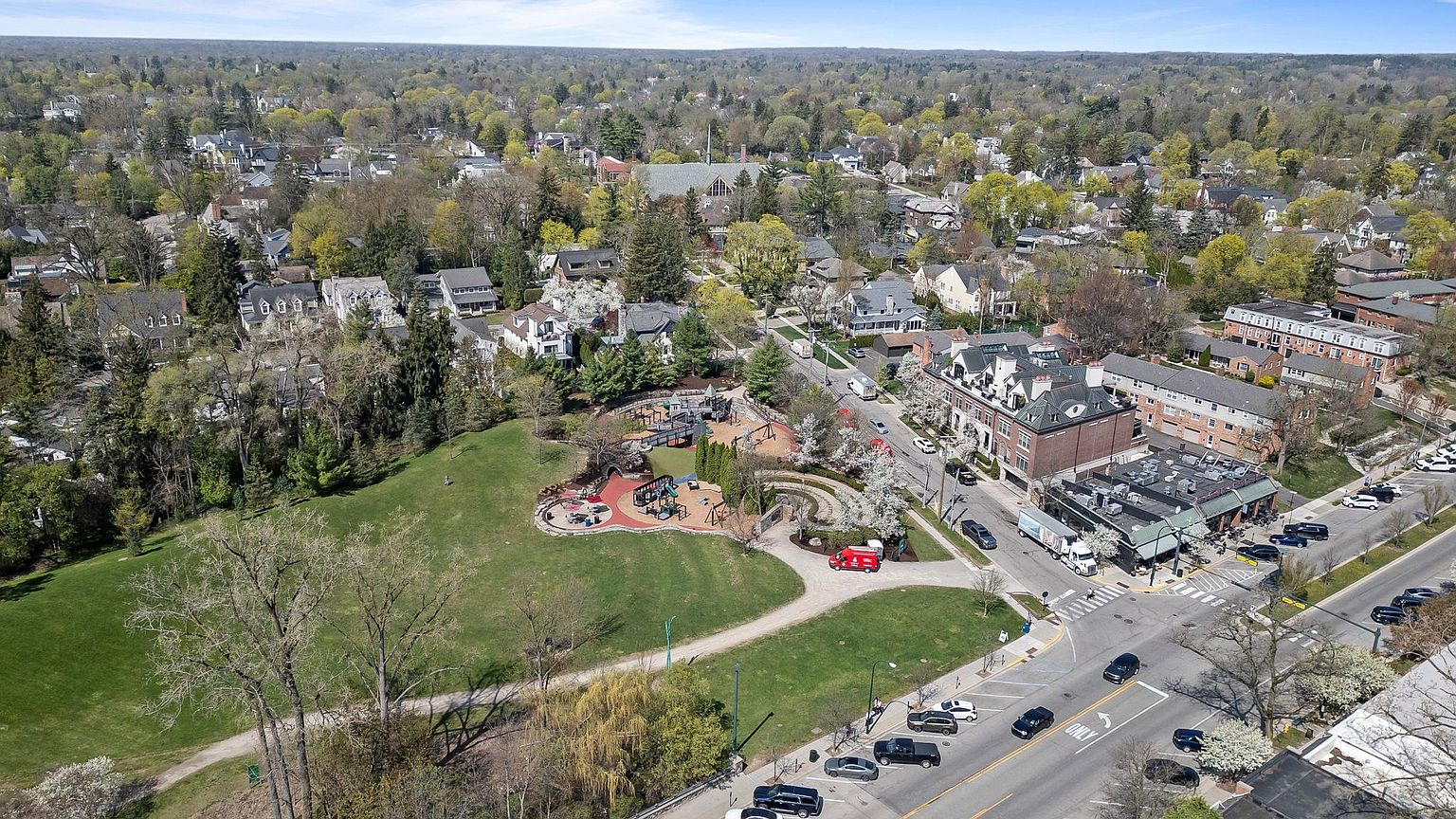 This aerial view showcases a charming neighborhood with a mix of residential and commercial buildings, centered around a well-maintained park and playground. The scene is lush with mature trees, giving a sense of established community and green space. A bustling street with visible traffic adds to the vibrant atmosphere of the area.