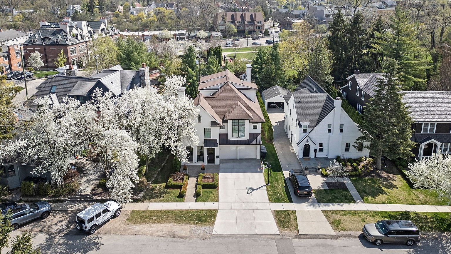 This aerial view showcases a well-manicured property featuring a modern two-story home with a brown roof and white exterior, complemented by mature trees and landscaping. A long driveway leads to a two-car garage, and several vehicles are parked on the street and in the driveway. The surrounding neighborhood is visible, adding context to the property's location.