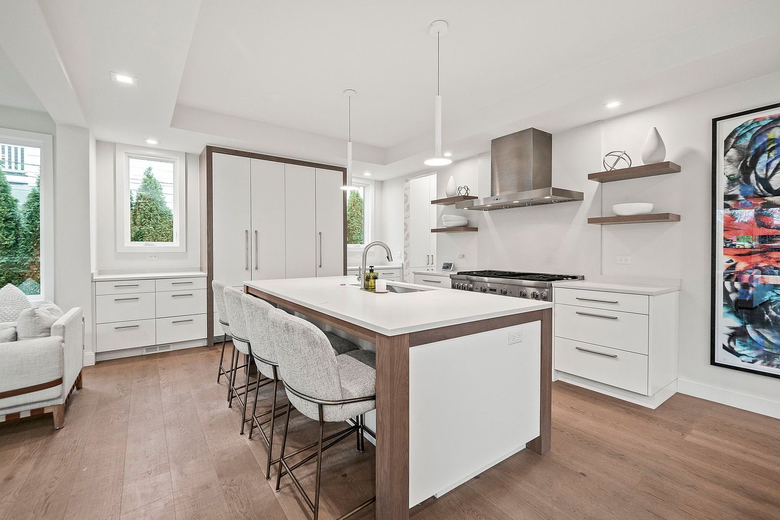 This is a bright and modern kitchen featuring a large island with seating, white cabinetry with dark trim, and stainless steel appliances. The kitchen is well-lit with recessed lighting and pendant lights over the island. Hardwood floors add warmth to the space, and a large piece of artwork adds a pop of color.