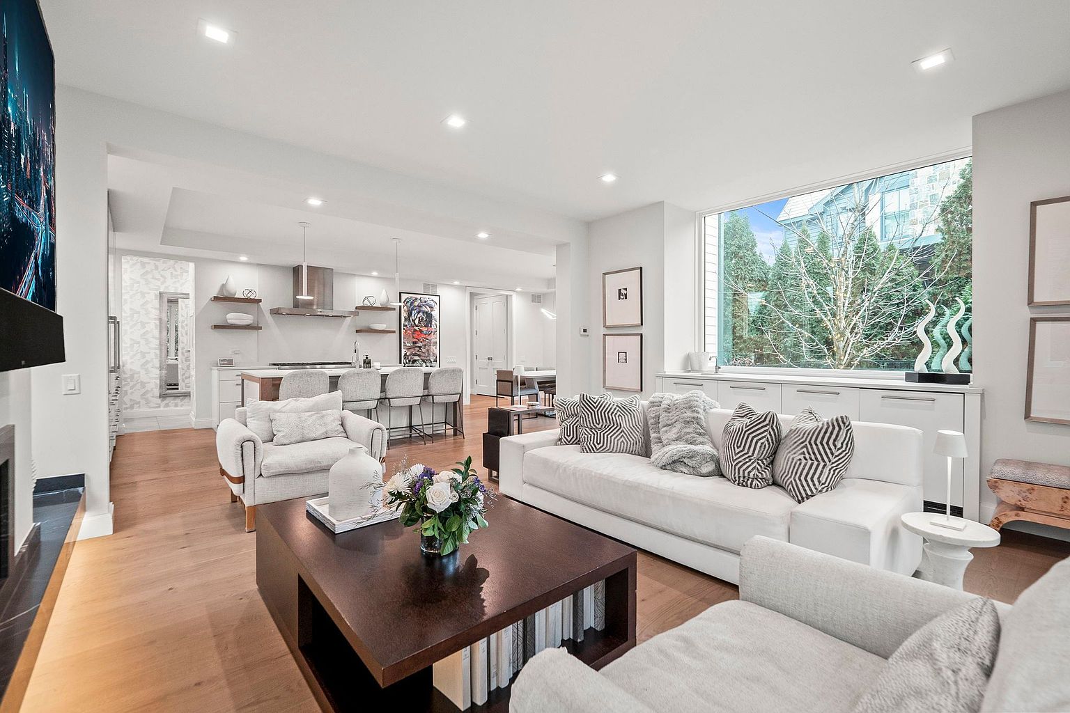 This is a bright and airy living room that seamlessly blends into an open-concept kitchen. The living room features hardwood floors, a large window providing natural light, and comfortable seating arrangements including a white sofa with decorative pillows and two armchairs. A dark wood coffee table with floral arrangements sits in the center, adding a touch of elegance.