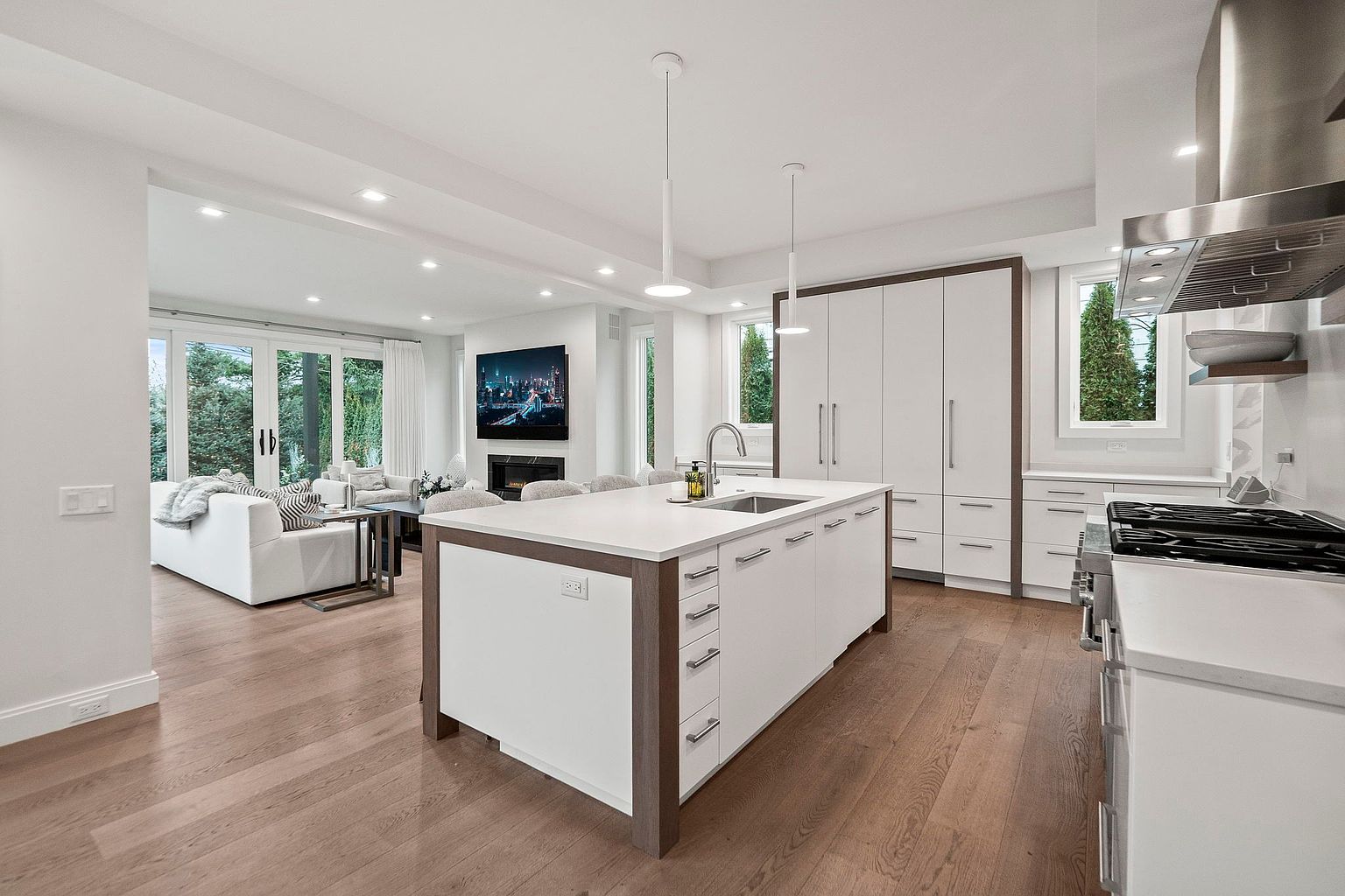 This is a bright, open-concept kitchen and living area. The kitchen features a large island with a sink, white cabinetry with dark trim, and stainless steel appliances. The living area has a white sofa, a fireplace with a TV above it, and large windows that let in plenty of natural light. The perspective is from the kitchen looking into the living area.