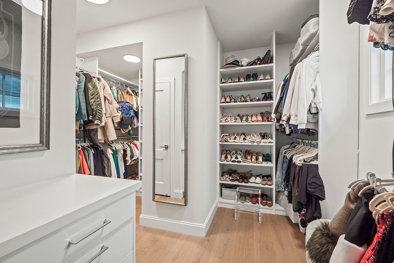 This is a well-organized walk-in closet featuring ample storage space for clothing and shoes. The closet includes hanging rods for clothes, shelving units for shoes, and a full-length mirror. The light wood flooring and white walls create a bright and clean aesthetic, enhancing the appeal of this organized space.