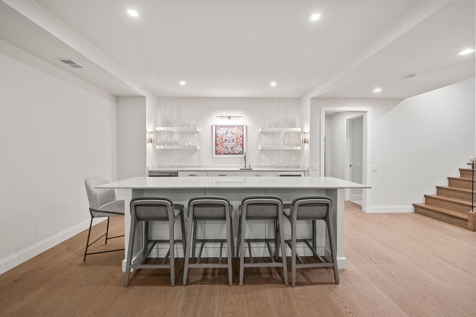 This is a well-lit basement area featuring a stylish bar with seating for four. The bar area includes white countertops, gray bar stools, and open shelving displaying glassware. The space is finished with light wood flooring and white walls, creating a clean and modern aesthetic, with a staircase visible on the right.