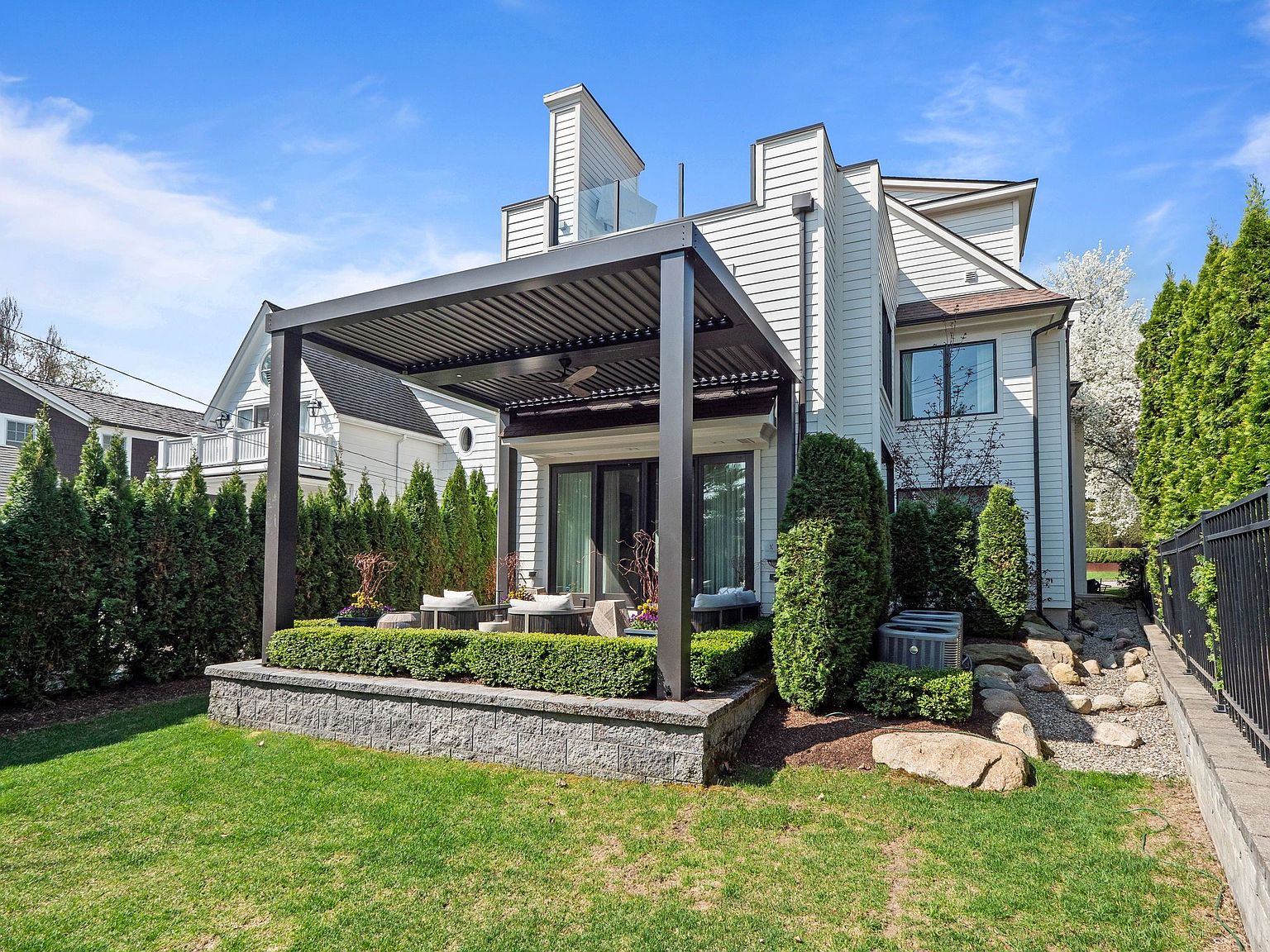 This image showcases a beautifully landscaped backyard featuring a modern pergola with outdoor seating. The yard includes a well-maintained lawn, manicured hedges, and a stone retaining wall. The house has white siding and a contemporary architectural style, creating an inviting outdoor living space.