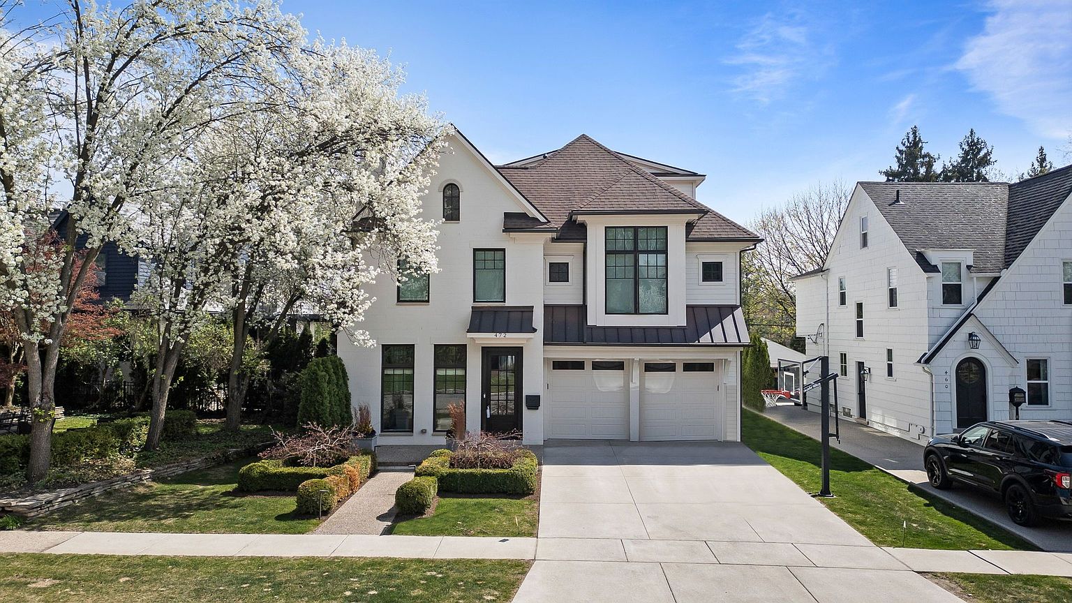 This is a front exterior view of a modern two-story house with a white painted brick facade, black framed windows, and a dark brown roof. The house features an attached two-car garage and a well-manicured front lawn with trimmed hedges and a walkway leading to the front door. A large flowering tree adds a touch of elegance to the landscape.