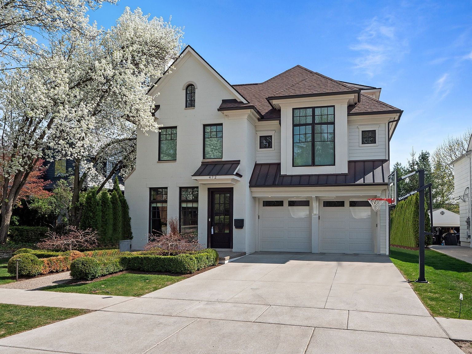 This is a front exterior view of a two-story modern home with a white brick facade and a brown roof. The house features black-framed windows, a two-car garage, and well-manicured landscaping with green bushes and a flowering tree. A basketball hoop stands on the right side of the driveway, adding a touch of recreational appeal.