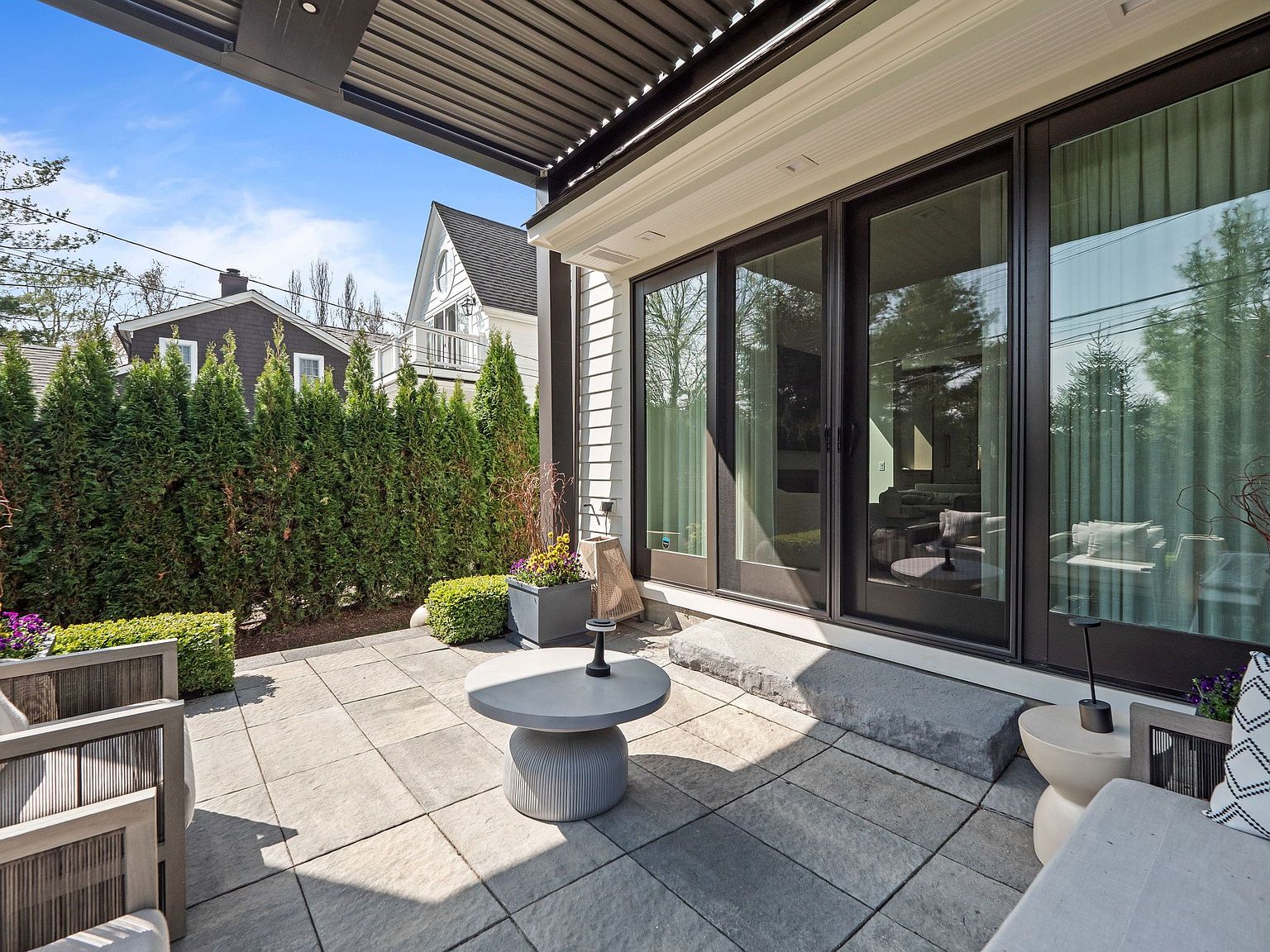 This image showcases a stylish outdoor patio area, featuring modern patio furniture, a round coffee table, and a well-maintained stone-tiled floor. Large sliding glass doors provide seamless access to the interior, while a lush green hedge offers privacy and a touch of nature. The patio is partially covered by a modern pergola, creating a comfortable and inviting outdoor living space.