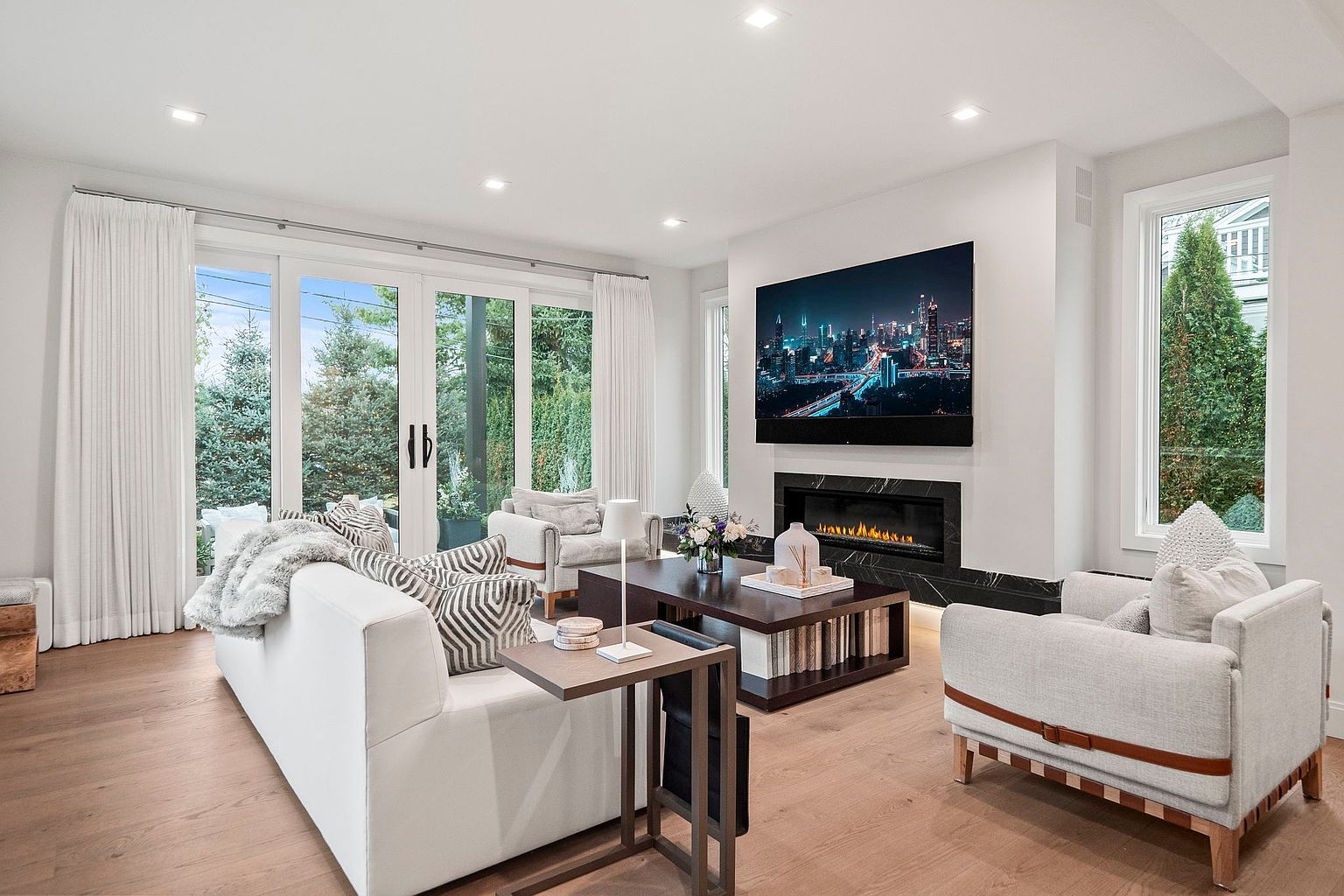 This is a bright and airy living room featuring a modern design with a large TV mounted above a fireplace. The room is furnished with a white sofa, light-colored armchairs, and a dark wood coffee table. Large sliding glass doors provide ample natural light and a view of the outdoors, enhancing the spacious feel of the room.