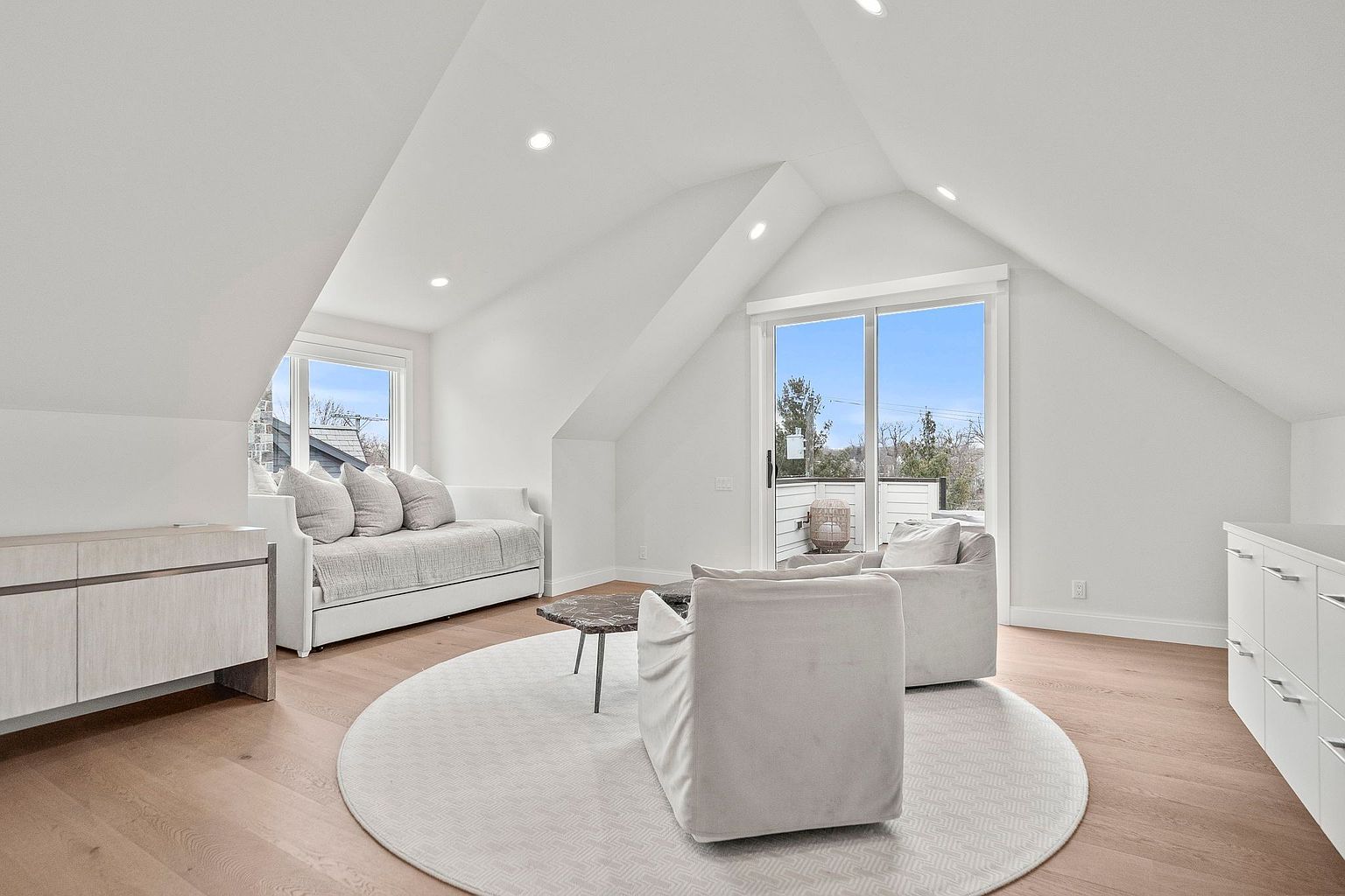 This is a bright and airy attic space converted into a cozy living area. The room features white walls, light wood flooring, and a round area rug. There is a daybed with pillows, two armchairs, a small coffee table, and built-in cabinetry, creating a comfortable and stylish retreat.